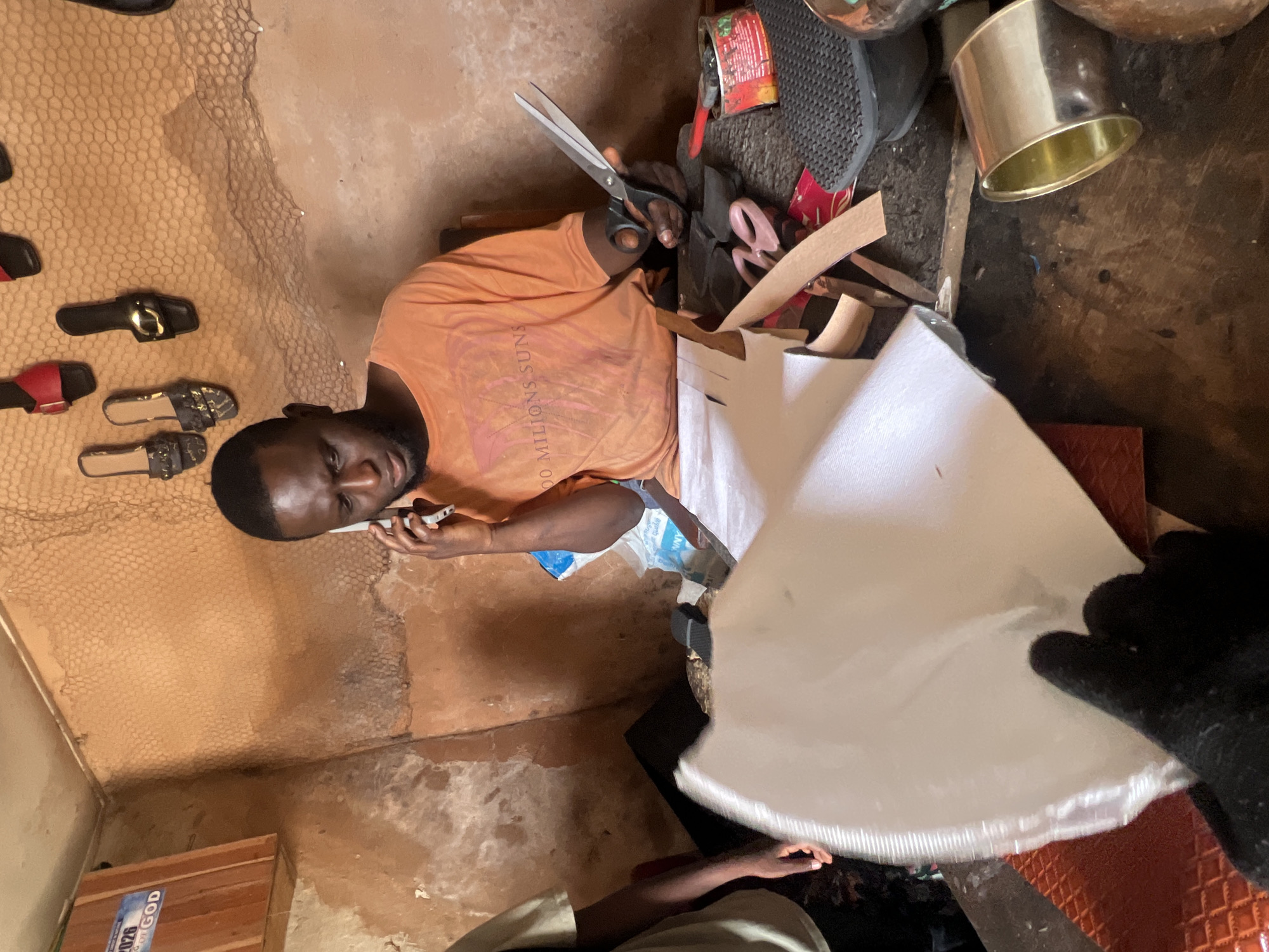 A dark-skinned man with short hair and a beard is sitting at a table in what appears to be a shoe-making workshop, possibly located in Jos, Nigeria. He is wearing an orange t-shirt with a faded graphic. He is holding a white sheet of material in his left hand, and in his right hand, he holds a large pair of scissors. The man is speaking on a silver mobile phone, which he holds to his left ear with his left hand.
The table is cluttered with various items, including rolls of brown leather, pink scissors, a red can, a black shoe sole, and a metal cylinder. The background shows a wall with a rough, textured surface, likely made of mud or clay. A net is stretched across the upper portion of the wall, and several pairs of sandals are hanging on it. To the left of the man, another person's arm is visible, wearing a light-colored t-shirt. On the wall to the far left, a wooden box with a sign that says "2026 God" is mounted. The overall environment suggests a humble workshop, possibly a small business or artisan's space. The lighting appears to be natural, coming from an unseen source.