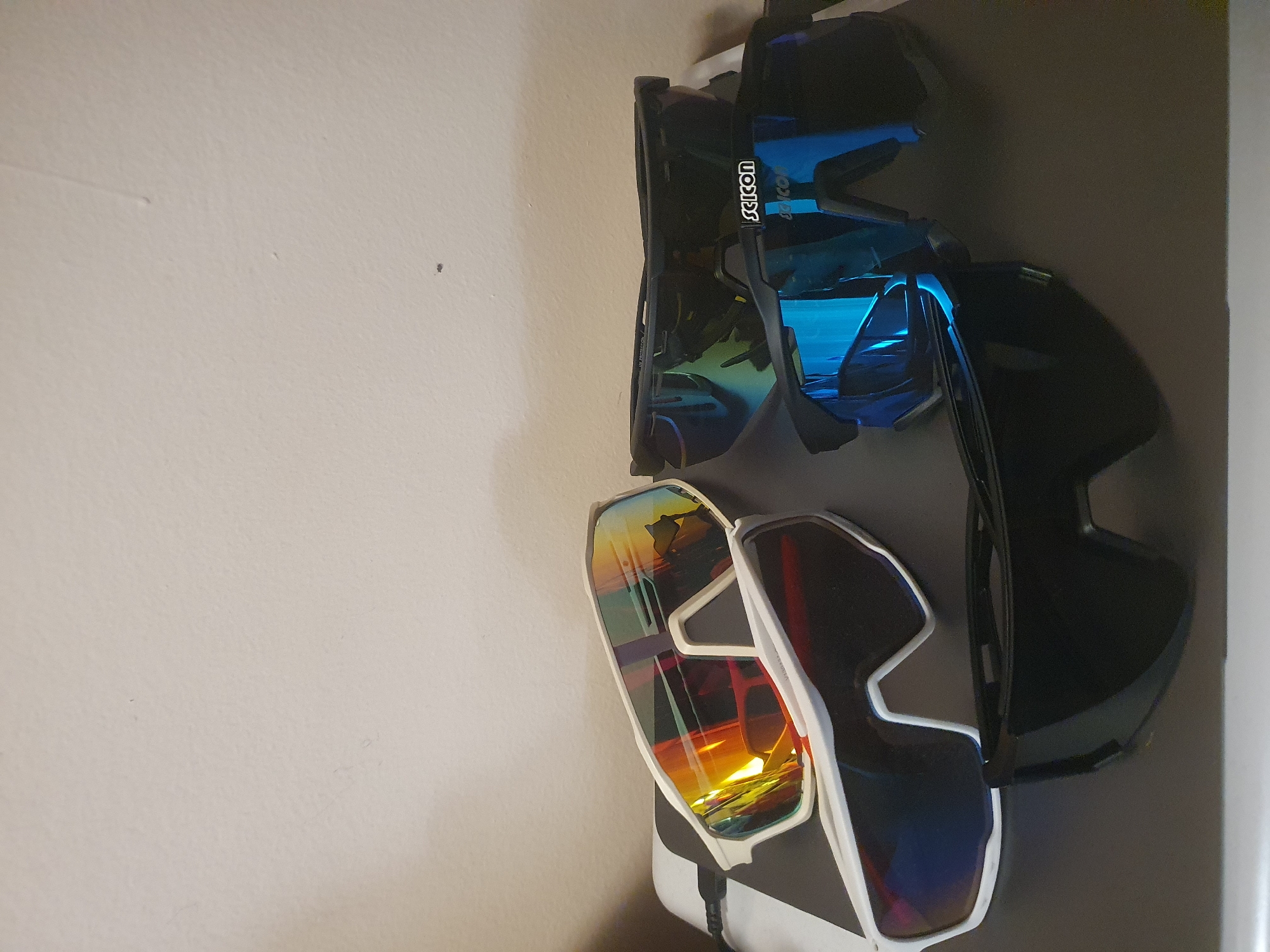 The image shows three pairs of sunglasses hanging on a wall. The top pair has blue lenses and the brand name "Scicon" visible on the frame. The middle pair appears to be a larger goggle-type frame with darker lenses. The bottom pair has white frames with iridescent lenses. The wall is light-colored and indistinct. There is no information in the image to suggest a location in Plovdiv, Bulgaria.