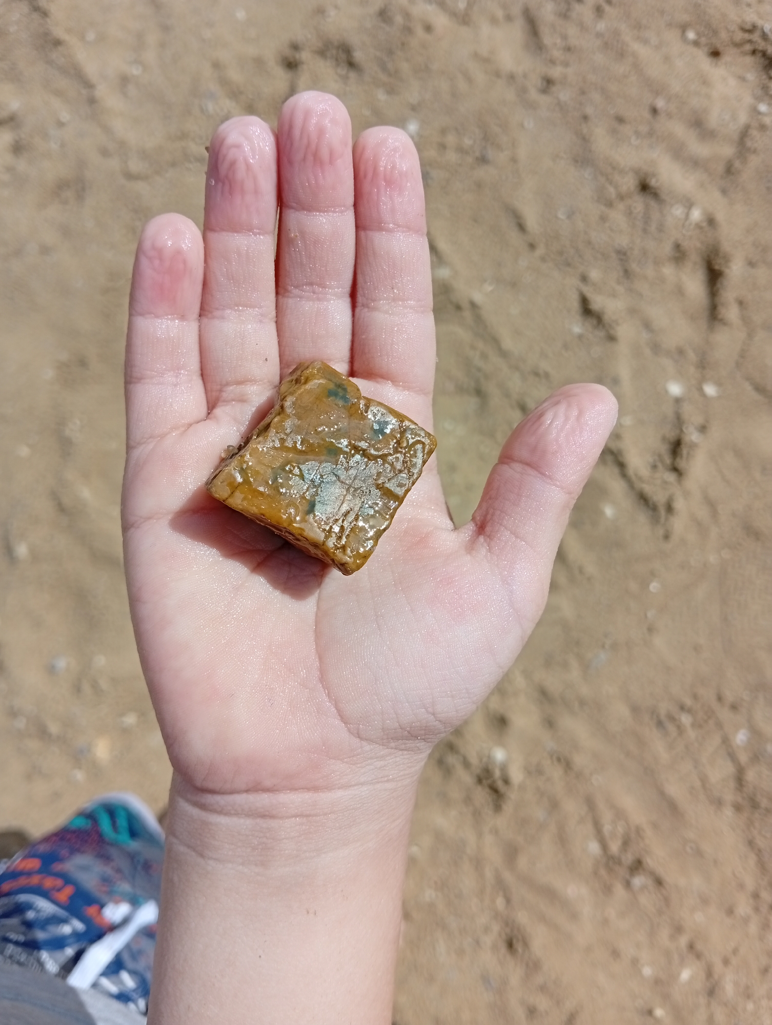 A person's hand holds a small, square-shaped, brownish-yellow rock with some light green or blue mineral inclusions. The rock is wet and appears slightly translucent. The hand is pale and damp, with visible finger creases. The background consists of dry, light brown sand, likely on a beach or a sandy area. The scene appears to be outdoors, possibly in a natural environment. No specific location within Мичурин ауылдық округі, Kazakhstan can be determined from the image alone.