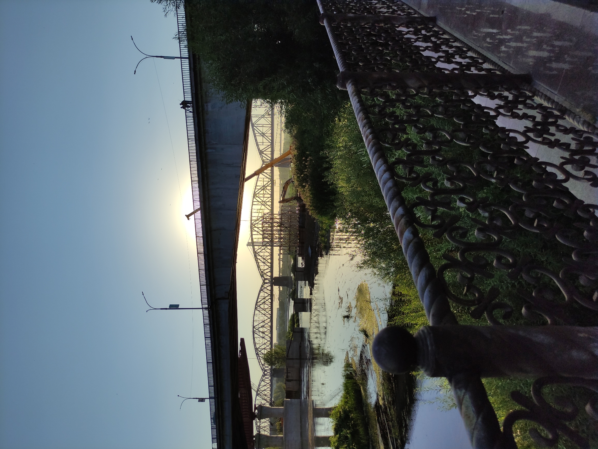 A wide-angle, eye-level shot captures a river with bridges in Semey, Kazakhstan at sunset. The sky is a pale blue, transitioning to a brighter yellow near the horizon where the sun is setting. Three bridges are visible. The closest bridge is a modern concrete and steel structure, with its underside illuminated by the setting sun. Beneath it, a more distant, older truss bridge with intricate metalwork spans the river. A third, even older bridge, also of truss design, is partially obscured by the middle bridge.
The river is calm, with reflections of the bridges and the sky on its surface. Algae and patches of green vegetation float on the water. Along the riverbanks, lush green trees and bushes are abundant. In the foreground, a decorative metal fence with a twisted rope-like railing borders a walkway. Two silhouetted figures stand on the topmost bridge, facing the sunset. Construction equipment, including a crane and an excavator, are visible near the middle bridge, suggesting ongoing work. The overall scene is serene and picturesque, with the warm glow of the sunset illuminating the bridges and the surrounding landscape.