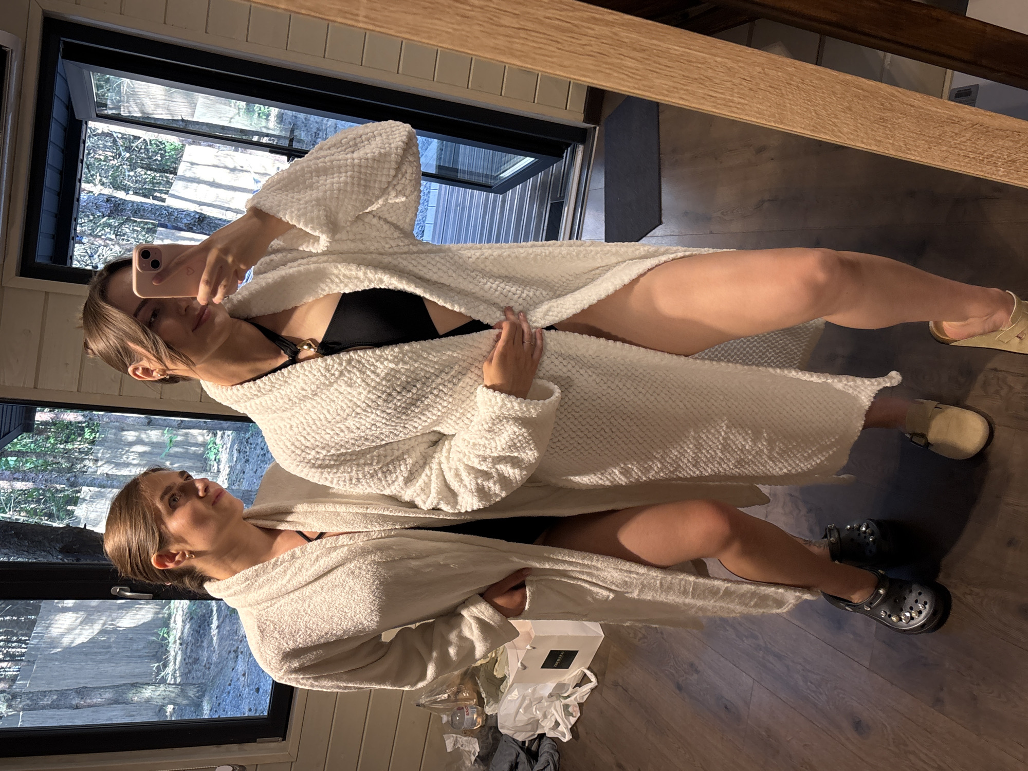 In the city of Ivachiv Horishnii, Ukraine, two women stand in a room with wooden paneled walls and a wooden floor. They are both wearing long white robes, and one woman is taking a selfie in a mirror. The room has a large window with a view of trees. There are several shopping bags on the floor near the women, along with a water bottle. One woman is wearing black Crocs with embellishments, and the other is wearing tan slip-on shoes. The scene appears to be in a private residence or accommodation.