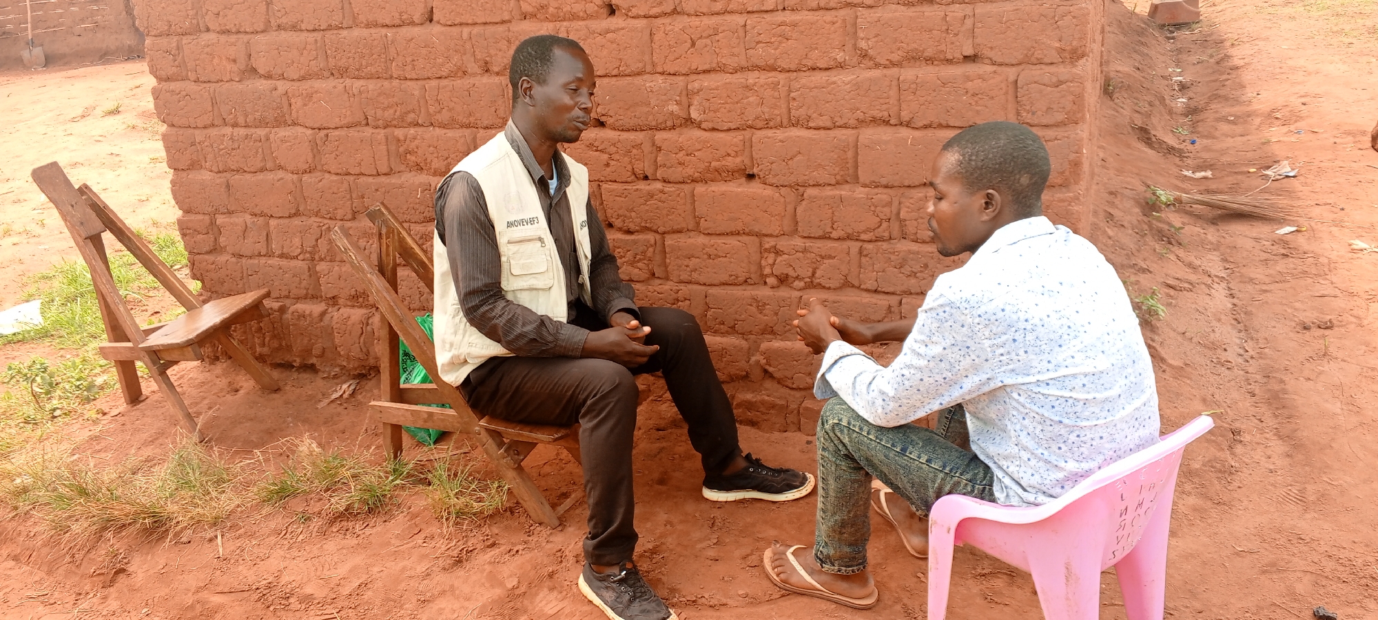 In an outdoor setting in Makungu, Democratic Republic of the Congo, two men are engaged in conversation. One man, dressed in a dark shirt, dark pants, and black shoes, is seated on a wooden chair. He wears a light-colored vest with the name "ANOVEVEE3" printed on it. The second man, wearing a light patterned shirt and jeans, sits on a pink plastic chair and wears flip-flops. The background includes a brick wall and reddish-brown dirt ground. Another wooden chair is visible, folded up, next to the seated man. A broom lies on the ground behind the man in the pink chair.