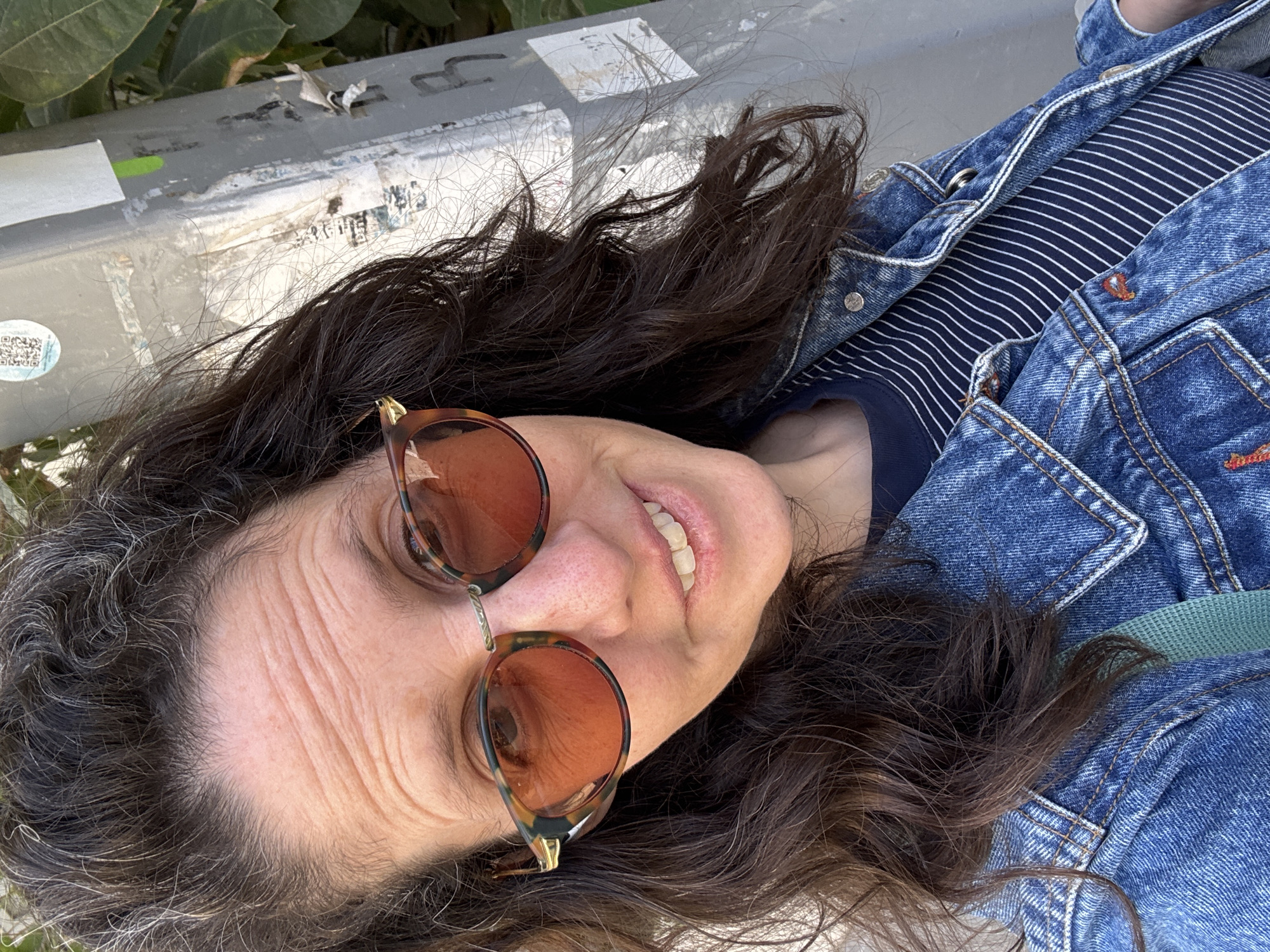 This medium close-up shot, taken in Tel-Aviv, Israel, features a person, likely an adult woman, smiling up at the camera. The image is rotated 90 degrees clockwise, giving a unique perspective as if the subject is looking up while lying down or from a low angle. Her head is positioned on a worn, grey, metallic surface, possibly a bench, railing, or wall, which shows signs of age, grime, and partially peeled stickers. Green foliage is visible at the top edge of the background.
The subject has long, dark brown, wavy hair mixed with significant strands of grey, especially around her hairline and temples. She wears tortoiseshell-framed sunglasses with amber lenses, which are casually perched on her nose rather than covering her eyes. Her forehead shows prominent wrinkles, suggesting an older individual. Her mouth is open in a natural, friendly smile, revealing her teeth.
She is dressed in a dark blue t-shirt with thin white horizontal stripes, worn beneath a classic blue denim jacket. The jacket features visible stitching, metal buttons, and pockets, consistent with a typical denim design. A green strap, possibly from a bag, is visible peeking out from under her jacket near her shoulder.
The setting is outdoors during bright daylight, indicated by the clear lighting and shadows. On the metallic surface behind her head, several distressed stickers are visible, including a small, round sticker containing a QR code on the left side, and various other illegible, faded markings and remnants of posters or stickers. The overall mood conveyed is relaxed and cheerful.
