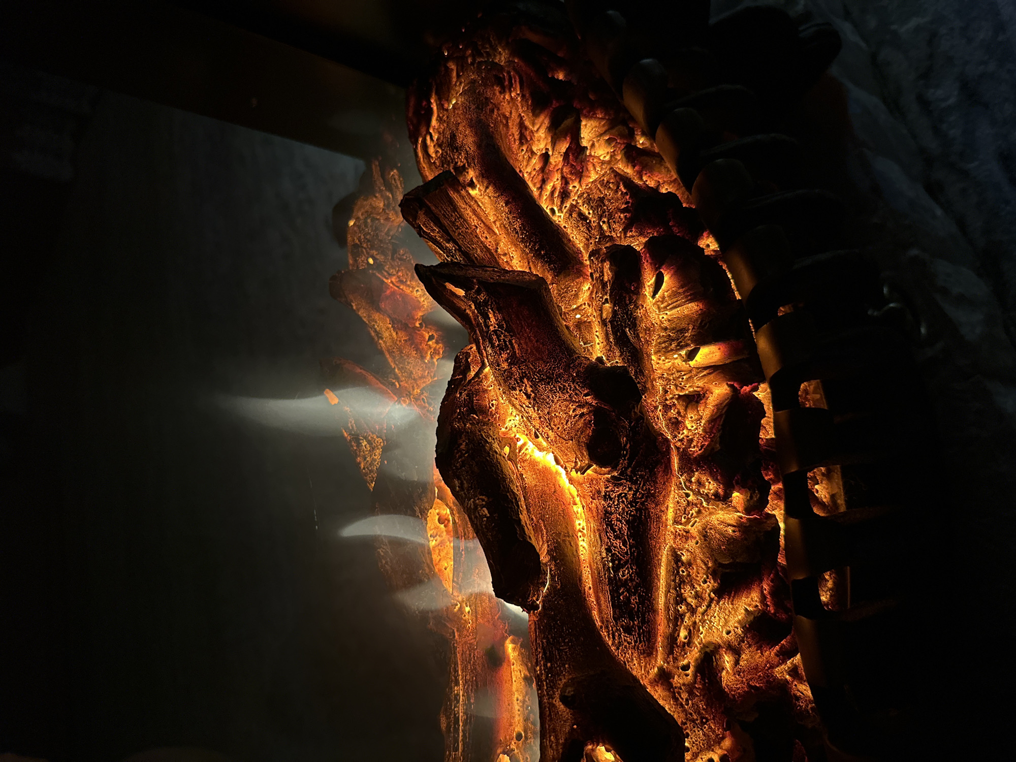 A close-up, dimly lit shot captures the glowing embers and simulated flames of an electric fireplace. The logs appear rough and textured, with vibrant oranges and yellows illuminating their surfaces. Wisps of white smoke or steam rise from the top of the logs, creating an illusion of heat. The background is dark and indistinct, with a hint of textured material, possibly a wall or surround, visible at the bottom. The overall impression is one of warmth and a cozy ambiance. The location is unknown.