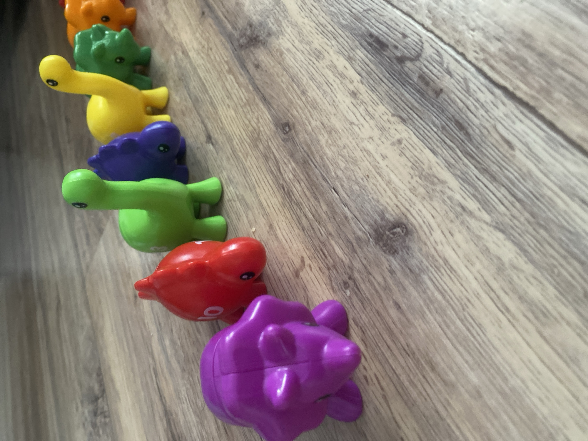The image shows a close-up, slightly angled view of a row of colorful, plastic dinosaur toys lined up on a wooden floor. The dinosaurs are arranged in a series: orange, green, yellow, purple, green, red, and purple. They are positioned on a light brown wooden floor with visible grain patterns. The location and city are Unknown.