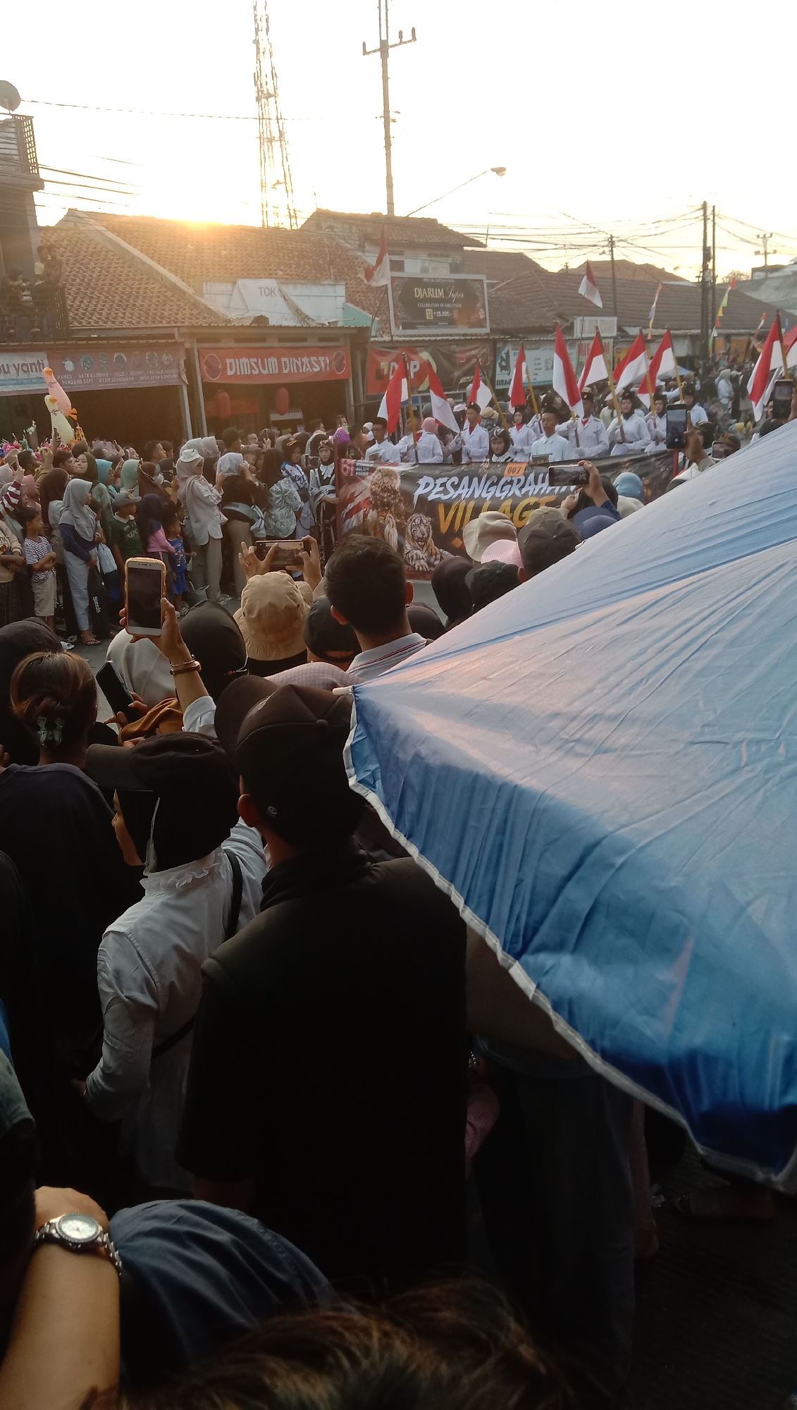 An outdoor street scene in Jatisawit, Indonesia, features a crowd gathered along a street lined with buildings. The sun is setting, casting a warm glow over the scene. In the foreground, a large blue umbrella partially obscures the view. A person in the foreground on the left is wearing a wristwatch with a silver band and a white face. Behind the umbrella, a group of people are visible, many holding up their phones to take pictures or videos. Some individuals are wearing white shirts and are holding red and white flags, indicating a possible celebration or parade. A banner with the text "PESANGGRAHAN VILLAGE" is also visible. The buildings in the background have red tile roofs, and there are some signs, including one that reads "DIMSUM DINASTI." A tall tower with antennas stands against the bright sky. The crowd appears to be watching or participating in an event.