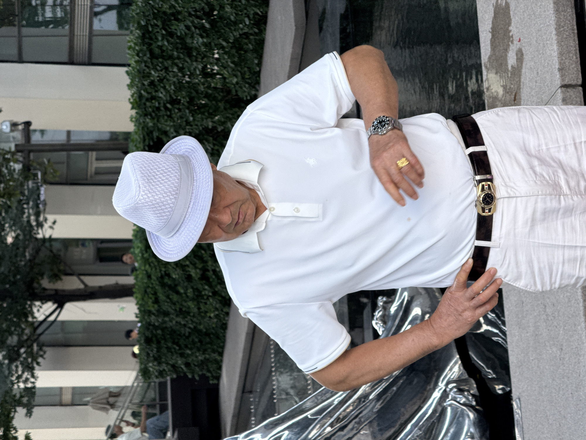 An older man wearing a white fedora, a white polo shirt, and white pants stands outside. He is looking down at his left hand, which has a gold ring on the ring finger. A watch is visible on his left wrist. He is also wearing a dark belt with a gold buckle. In the background, there is a green hedge, a building with large windows, and some trees. The man is standing next to a body of water and a shiny, abstract sculpture. The scene appears to be an urban outdoor setting.
