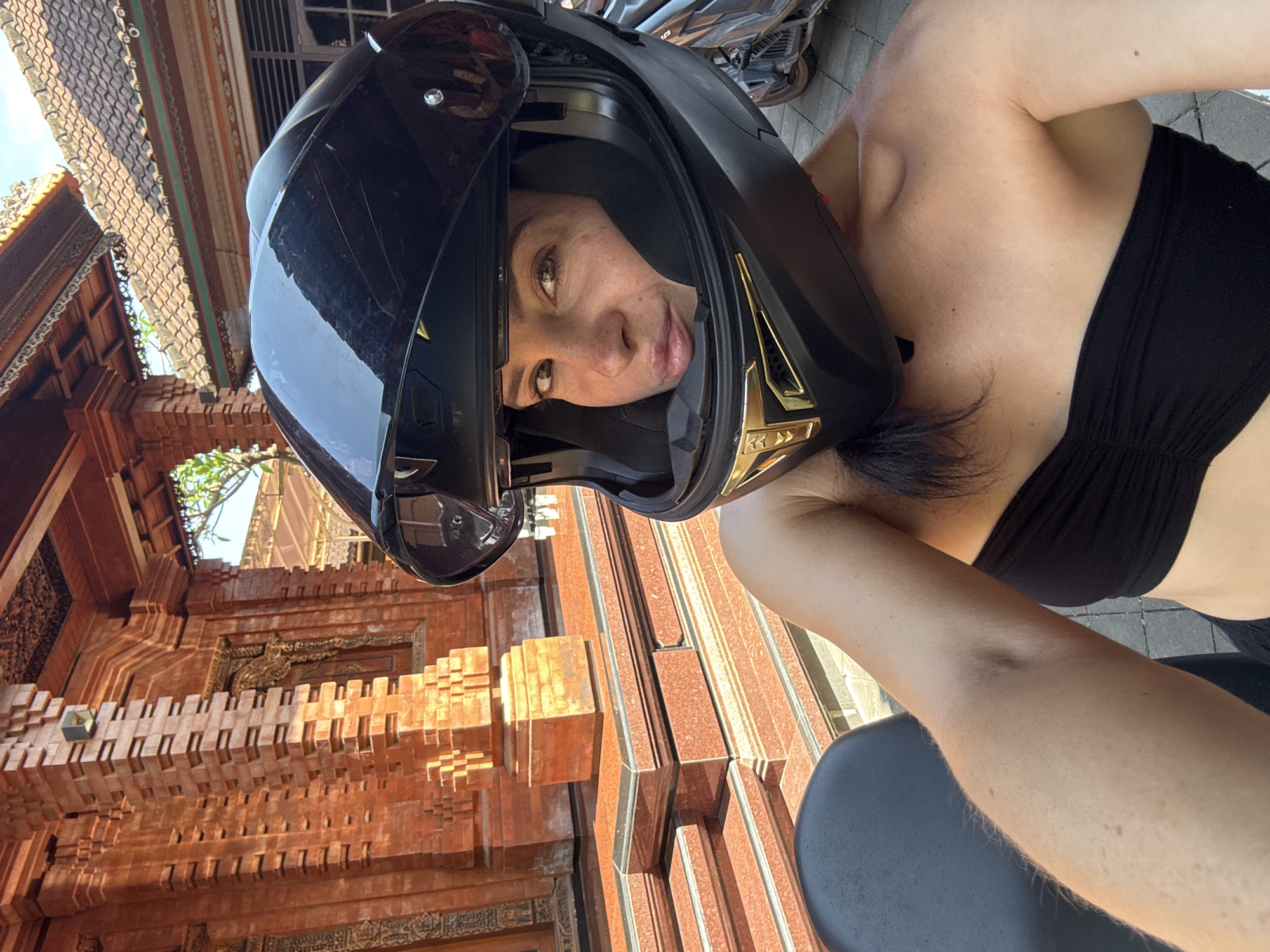 A woman, wearing a black motorcycle helmet and a black strapless top, is taking a selfie. She is looking at the camera with a relaxed expression. The background shows a traditional Balinese building with intricate brickwork and carved details, suggesting a location in North Kuta, Indonesia. The architecture, with its reddish-brown tones and ornate patterns, is characteristic of Indonesian temples or traditional homes. Sunlight is visible, indicating it is daytime, and the overall atmosphere seems casual and possibly adventurous, given the helmet and the implied mode of transportation. There is no visible text in the image.