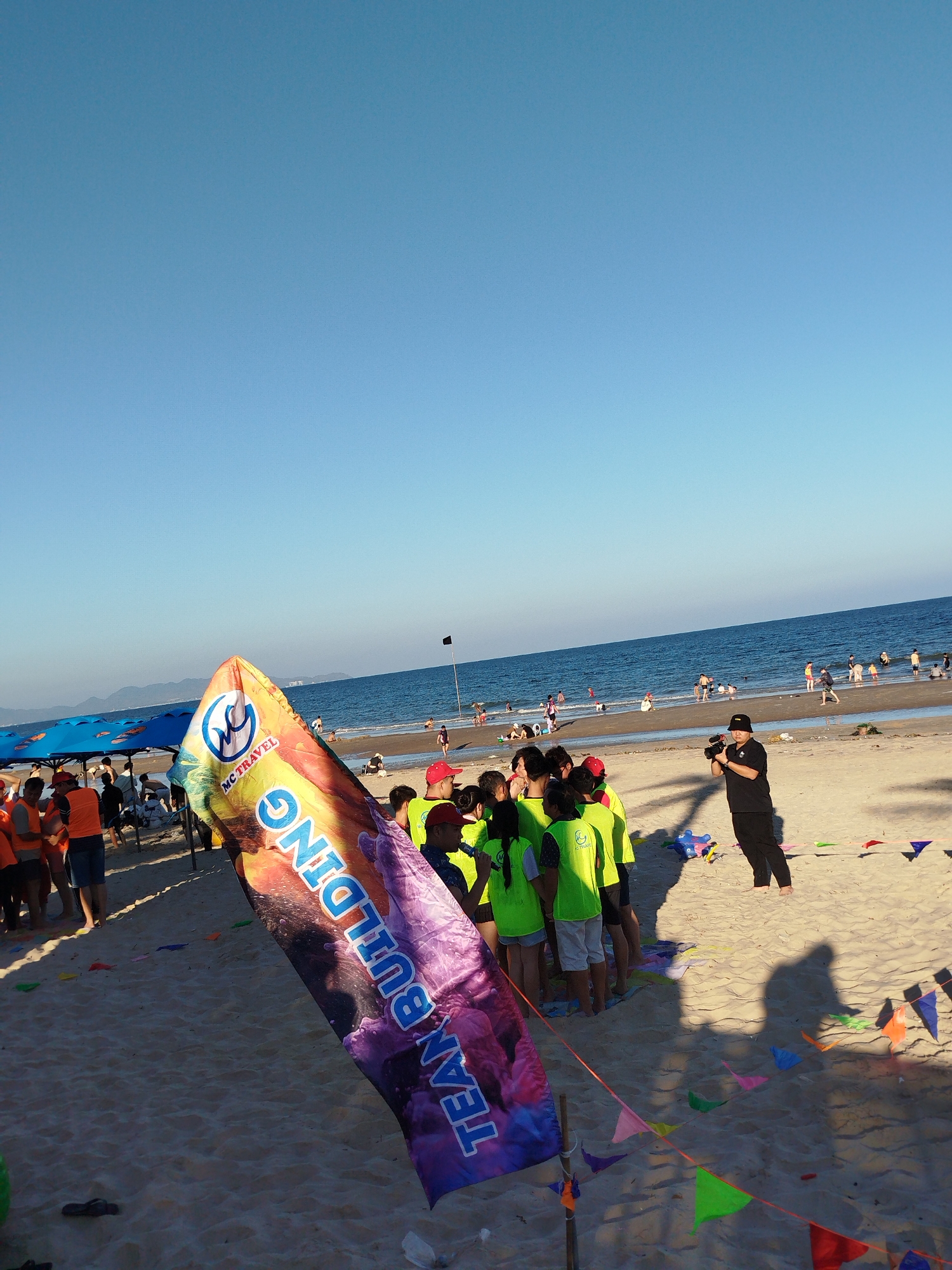 The image shows a sunny beach scene. A group of people wearing lime green shirts are huddled together, partially obscured by a large banner that reads "TEAM BUILDING" with an "MC Travel" logo on top. A man in all black, holding a camera, stands to the right of the group. In the background, the ocean meets the horizon under a clear blue sky, with a scattering of people on the beach and in the water. Small triangular flags are strung along a rope near the foreground.
The specific location of the image cannot be determined as Ho Chi Minh City does not have a coastline. While it is possible that the people are from Ho Chi Minh City, the exact geographic location is indeterminate.