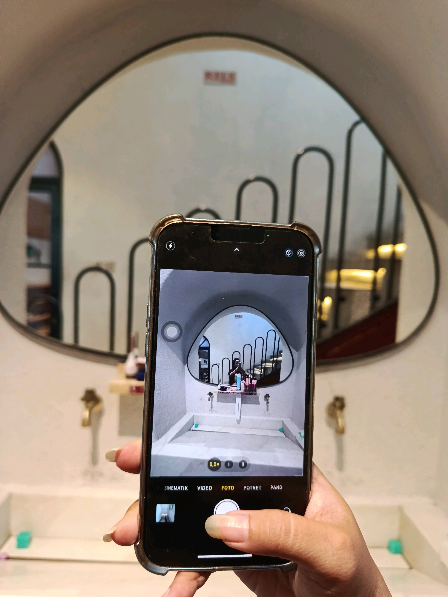 A hand holding a smartphone is in the foreground, capturing a photograph of a bathroom with a large, heart-shaped mirror. The smartphone screen displays the reflection in the mirror, showing a sink area with faucets and toiletries. The background of the mirror shows a staircase with decorative metal railings and illuminated steps. The bathroom itself is minimalist, with white sinks and tiled walls. There are no discernible street signs or landmarks to indicate the specific location within Kudus, Indonesia. The lighting is warm and ambient, suggesting an indoor setting.