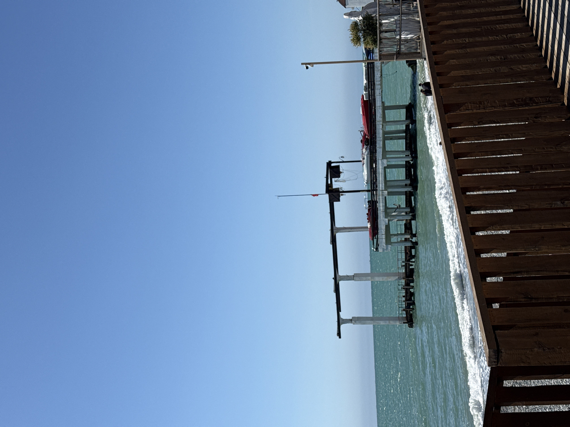 This image depicts a sunny day at a pier in Sochi, Russia, offering a clear view of the sea and coastal structures under a cloudless blue sky.
On the right side of the frame, a robust wooden walkway or pier extends diagonally from the lower right towards the upper-middle. Its distinct planks and strong shadows indicate bright, direct sunlight. To the left of this wooden structure, the turquoise-green sea is visible, with gentle white-capped waves breaking along the edge.
Further into the sea, a more elaborate pier structure is supported by several light-colored concrete pillars. This structure features horizontal platforms and railings, and several boats or watercraft are docked alongside it, including a noticeable red vessel. A tall pole within this structure holds a security camera, and other equipment, possibly fishing rods, can be seen. In the distant background, beyond the pier, glimpses of buildings and green foliage suggest the coastline.
The time of day appears to be mid-day due to the bright illumination and sharp shadows. The weather is clear and sunny, contributing to a calm and pleasant atmosphere. No people are visible in the scene, and there is no discernible text. The image captures a serene coastal environment typical of a resort city like Sochi.