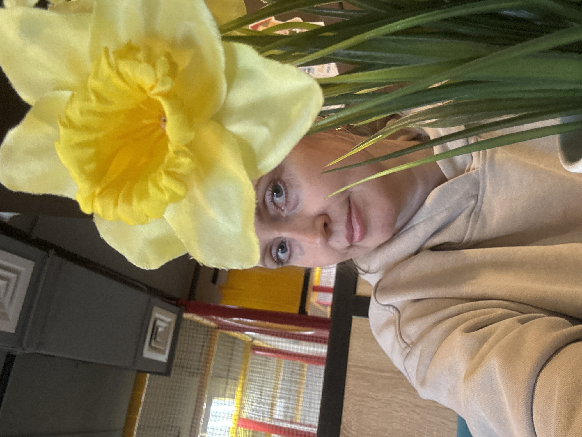 The image shows a person wearing a light-colored hoodie, positioned with a yellow flower in the foreground. Green plants surround the subject's head. In the background, a gray structure with decorative elements is visible, along with a colorful indoor playground with red and yellow elements. The location appears to be an indoor setting. The subject appears to be located in Chaikovsky, Russia.
