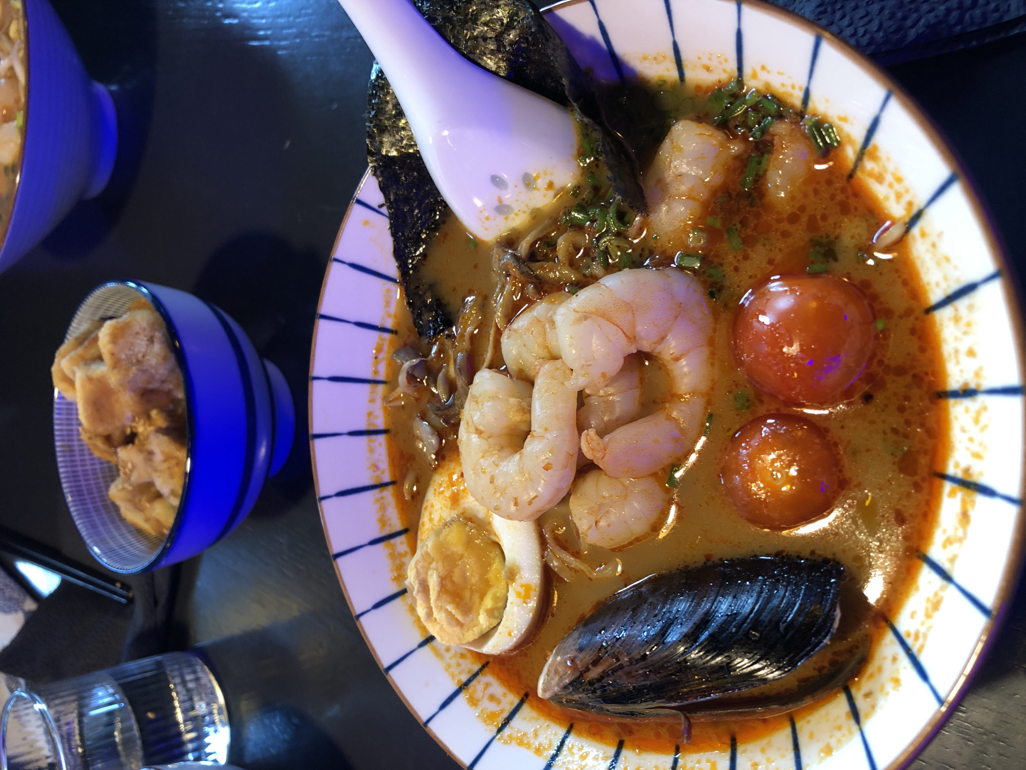 A close-up shot shows a bowl of ramen soup, a common dish in Japanese cuisine, served in a white bowl with blue stripes. The ramen is filled with various ingredients, including plump shrimp, a halved hard-boiled egg, cherry tomatoes, a black mussel shell, and dark green seaweed. The broth is a vibrant orange-red, suggesting a spicy flavor. A white spoon is partially submerged in the soup, indicating that someone is about to eat or has just taken a bite.
To the left of the ramen bowl, there is a small blue bowl filled with fried croutons or pieces of fried bread. A set of chopsticks rests beside the blue bowl. In the foreground, a clear glass with water is visible, and another glass is partially obscured. The table appears to be dark and reflective, possibly made of wood or a similar material. The lighting in the scene is dim and atmospheric, with a prominent blue light reflecting on the spoon and broth, suggesting an indoor setting perhaps at night. The overall impression is that of a casual meal being enjoyed. The image does not contain any visible text. The location context provided is Izhevsk, Russia.