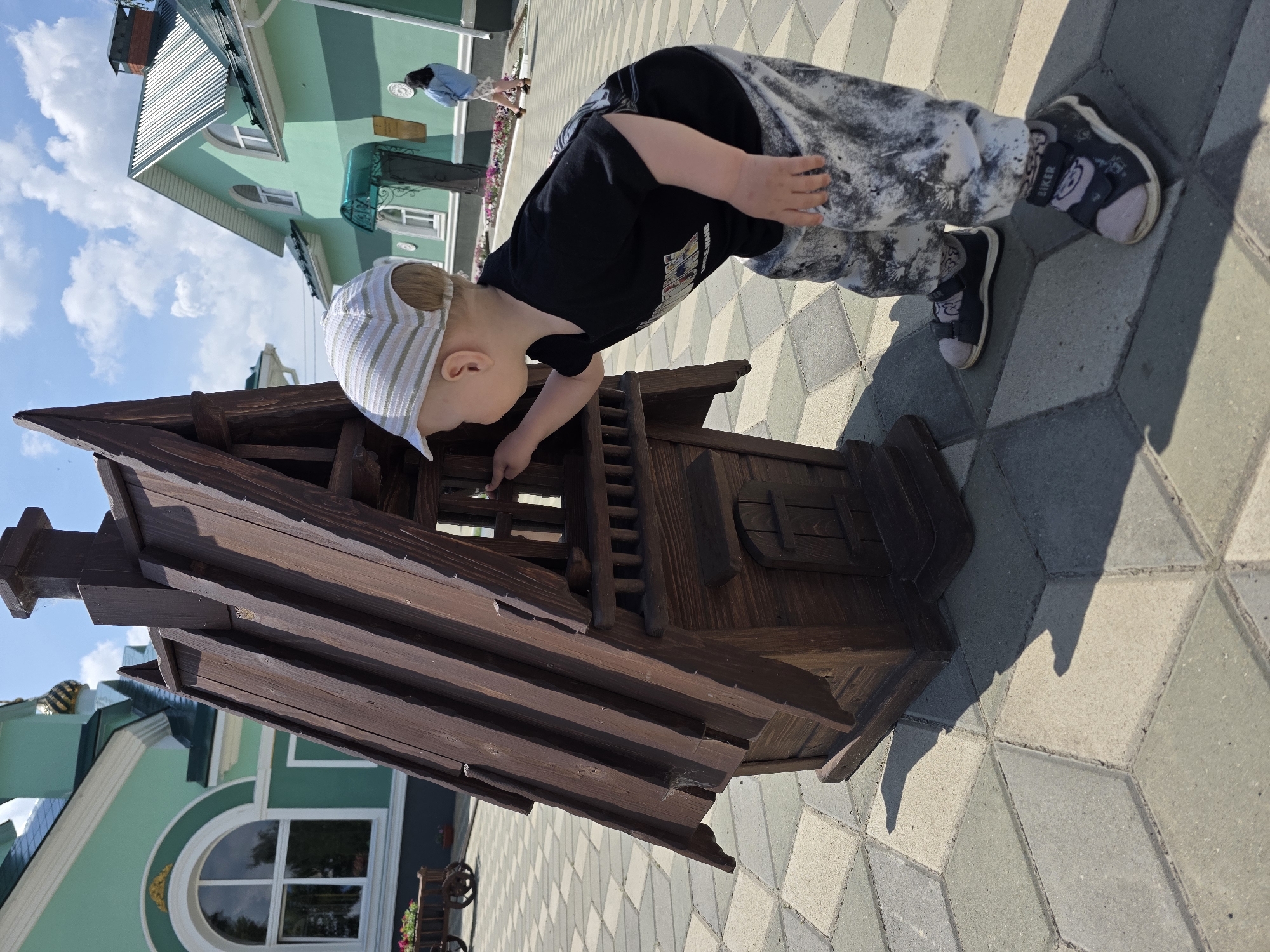 A young child wearing a black t-shirt, camouflage pants, and a striped cap is reaching into a wooden birdhouse on a paved area. The birdhouse is dark brown and has an angled roof. In the background, there are green buildings with white trim and arched windows. The sky is blue with white clouds. A woman in a blue jacket is visible in the distance. The scene appears to be a park or a public outdoor space in the city of Chaikovsky, Russia.