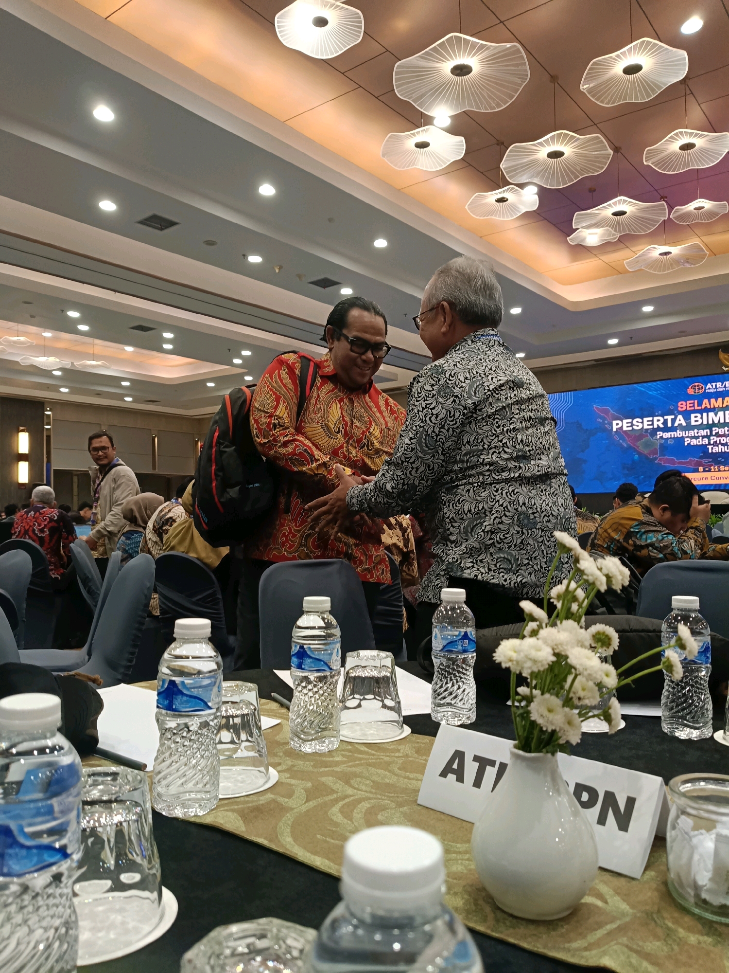 In a well-lit conference hall, two men are shaking hands. The man on the left, wearing a red and gold patterned shirt and glasses, appears to be smiling. The man on the right, in a black and white patterned shirt, is also looking at the man on the left. Both men have their right hands clasped. Behind them, a large screen displays text in Indonesian, including the words "SELAMAT" and "PESERTA BIMBINGAN". The screen shows a map of Indonesia. In the foreground, a table is set with water bottles, glasses, and a vase of white flowers. Several rows of empty chairs are visible in the background, with other attendees seated at tables. The ceiling features modern, cloud-like light fixtures. The scene suggests a formal event or conference, possibly in North Jakarta, Indonesia, given the Indonesian language on the screen.