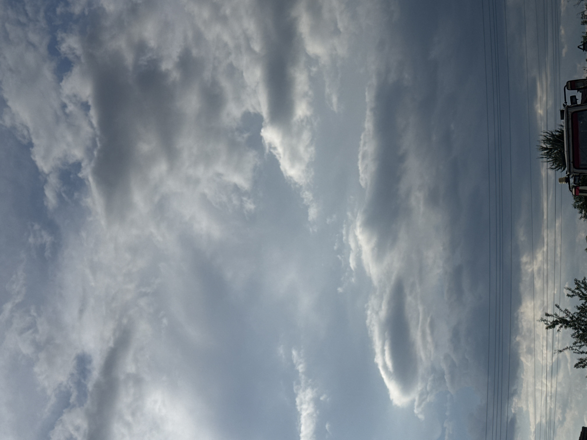 The image presents a daytime sky filled with a variety of cloud formations, exhibiting both thin wispy clouds and larger, more substantial cumulus clouds. A few powerlines are visible in the right side of the frame. A blurry, seemingly stationary vehicle is seen in the far right side of the frame, and some foliage is near it. I do not have sufficient information to determine whether the image was taken in Sarnevo, Bulgaria.
