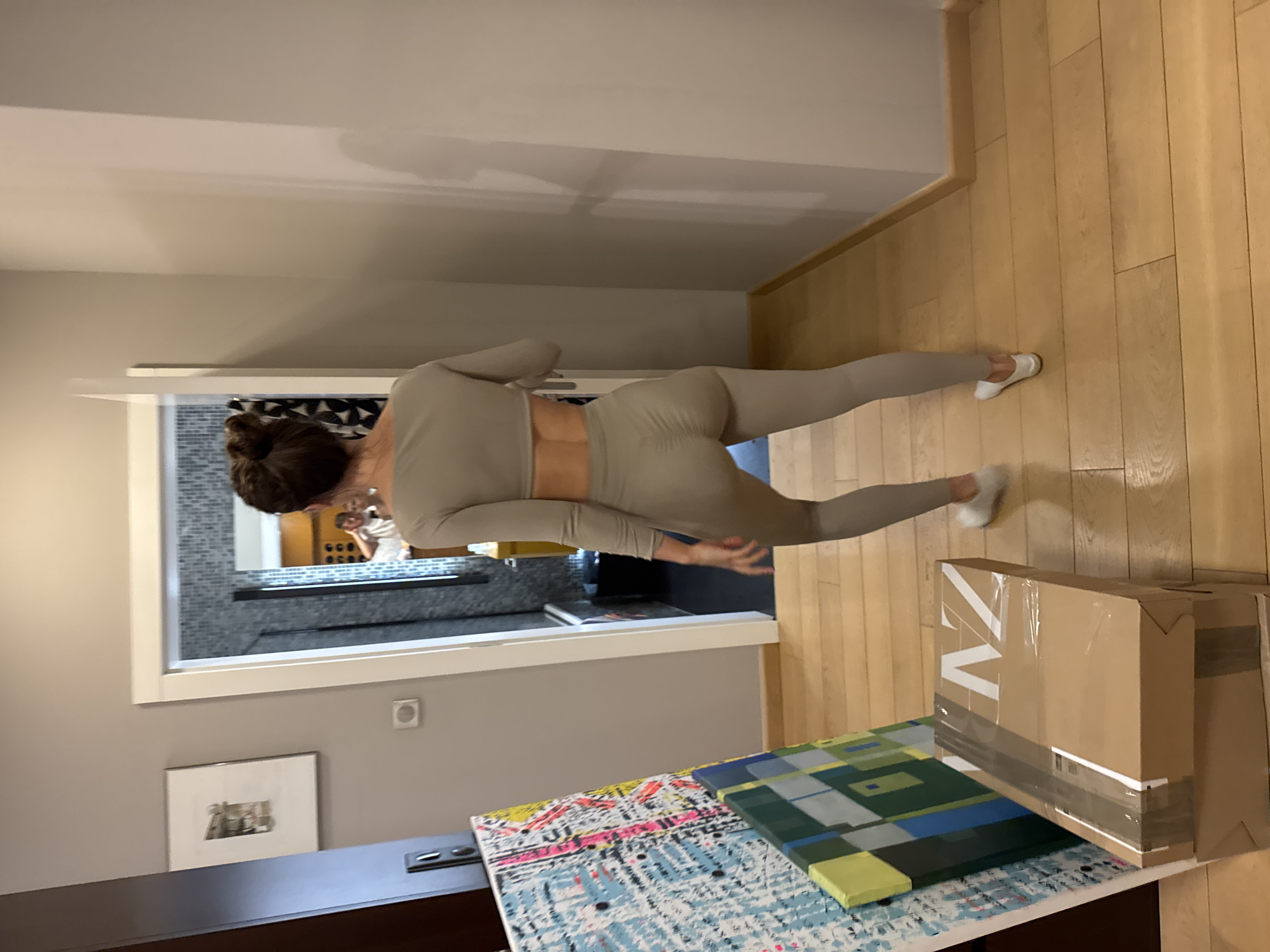 A woman in light-colored athletic wear stands in a hallway. The hallway has light gray walls and wooden floors. She is facing away from the camera, looking into a bathroom with a gray tiled wall and a mirror. Behind her, there is a box with white letters and what appears to be art canvas with geometric patterns on a table. A framed picture hangs on the wall of the hallway. The location is likely in Warsaw, Poland, based on the visual characteristics of the architecture.