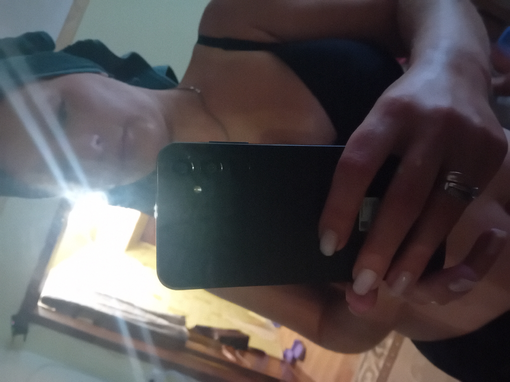 A person is taking a mirror selfie indoors. The person is wearing a dark bra and a teal towel wrapped around their head. They are holding a black smartphone in their right hand, pointed towards the mirror. The background shows a doorway with sunlight streaming through, and some wooden furniture. The floor has a patterned rug and a pair of purple flip-flops. The scene appears to be in a residential setting in Chuhuiv, Ukraine, likely a bathroom or bedroom.