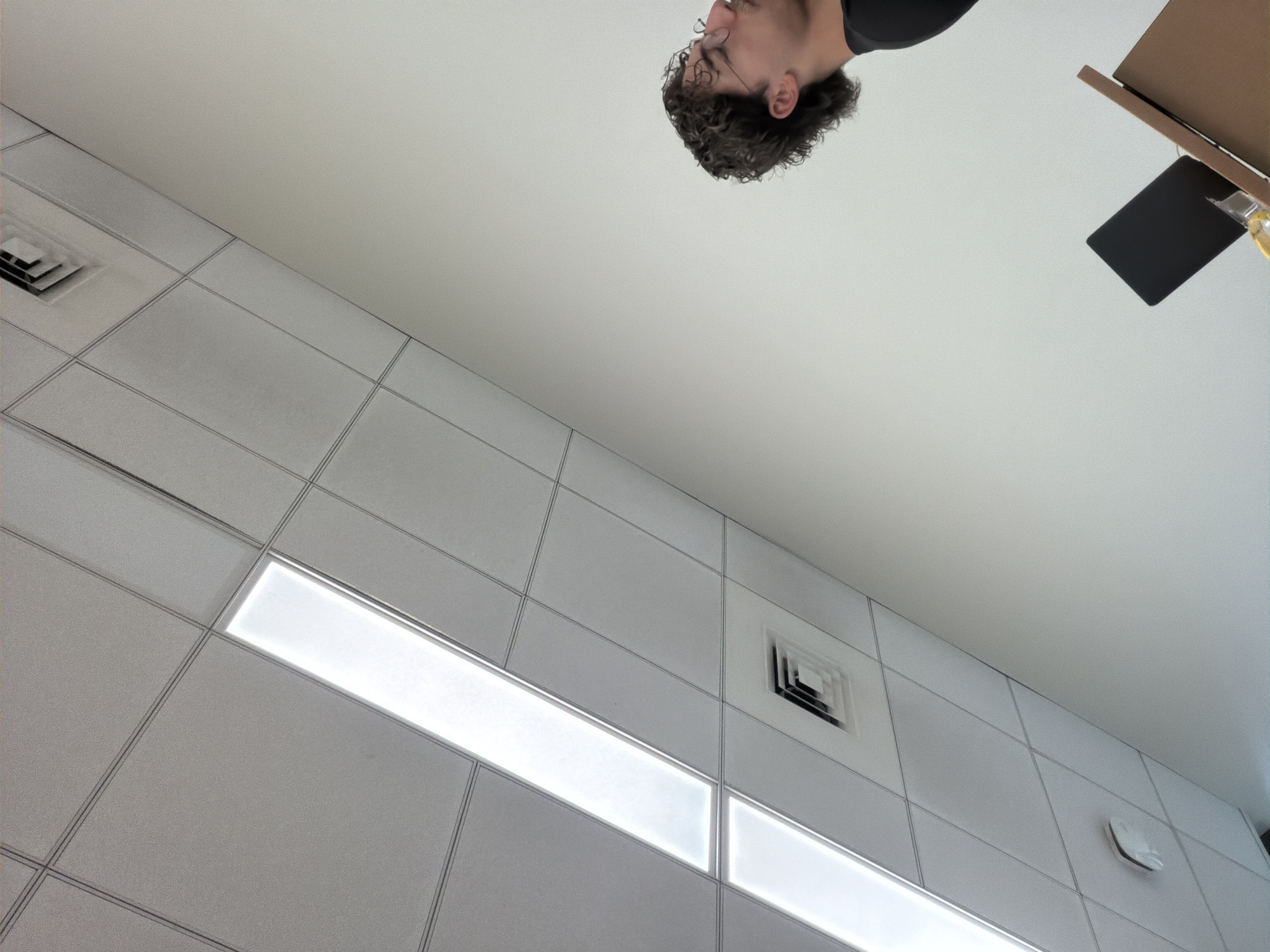 The image presents a low-angle view, likely taken from a ceiling, showcasing a person's head in the upper portion of the frame along with various architectural and technological elements. The person appears to have curly, dark hair and is wearing a black top. Next to them, at their right, there are items suggesting a tablet, a cardboard box-like structure, and what could be a bottle. The background primarily consists of a modular ceiling structure, with square tiles and linear lighting fixtures. Additionally, there are visible air vents integrated into the ceiling grid. The scene has a contemporary and somewhat sterile feel, resembling the interior of an office or public building. There is no indication of the city of New York within the image. The actions and interactions are difficult to ascertain due to the nature of the photo.