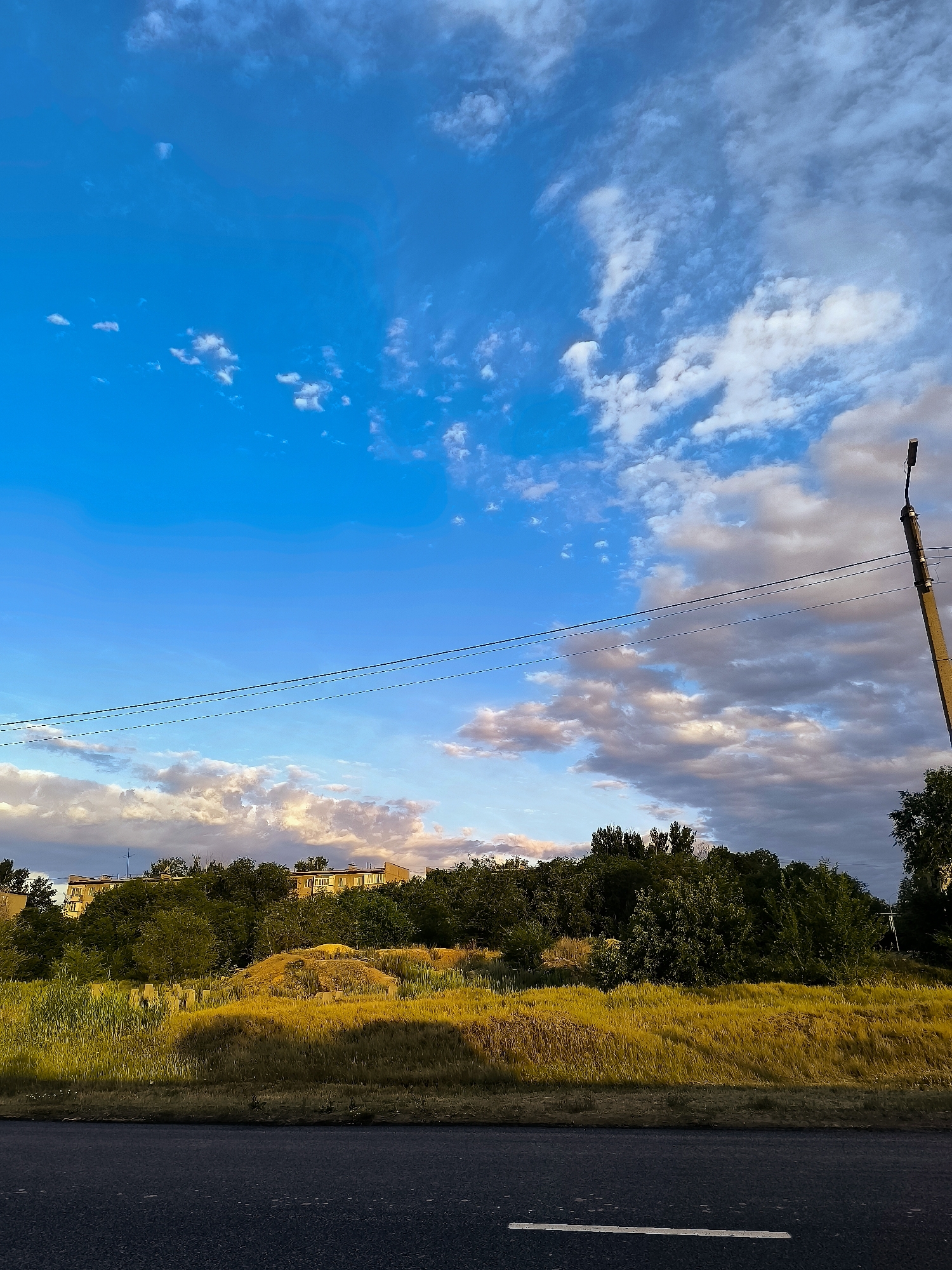 The image depicts a bright, overcast sky with a mix of blue sky and scattered white and gray clouds, suggesting a potentially windy day. The sun is shining through, casting a warm, golden light on the vegetation in the foreground. The scene appears to be a roadside with a dark asphalt road in the lower part of the frame, marked by a white line.
Behind the road, there is a slope covered in tall, dry, golden grass. Further back, a dense line of green trees forms a natural barrier, behind which a multi-story building with light-colored facades is visible. The building has a few windows and appears to be an apartment complex or a similar structure. On the right side of the image, a tall, rustic utility pole with wires extending from it stands out against the sky. The overall impression is one of a rural or semi-urban setting on a bright, possibly late afternoon, given the quality of light. There are no people visible in the image, and the mood is serene and natural. The text "Qarağandı city administration, Kazakhstan" provides the geographical context for this image.