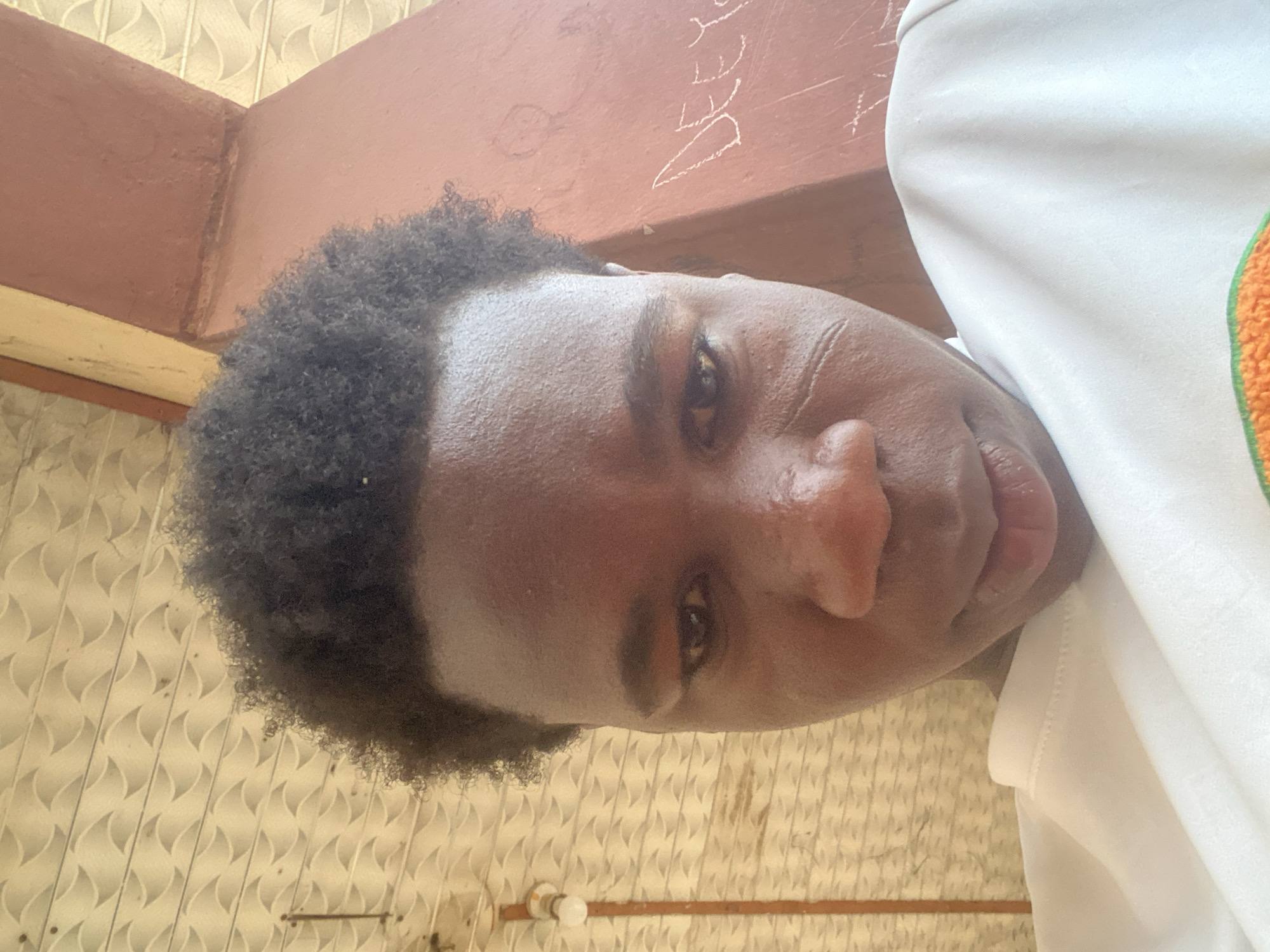 A close-up shot looking down at an individual with dark skin and short, curly hair. The person is wearing a white shirt with a visible orange trim on the shoulder. The background includes a brown structure with some white writing on it and a wall with a patterned tile design. The image was likely taken indoors in Tudun Wada Bibinu, Nigeria.