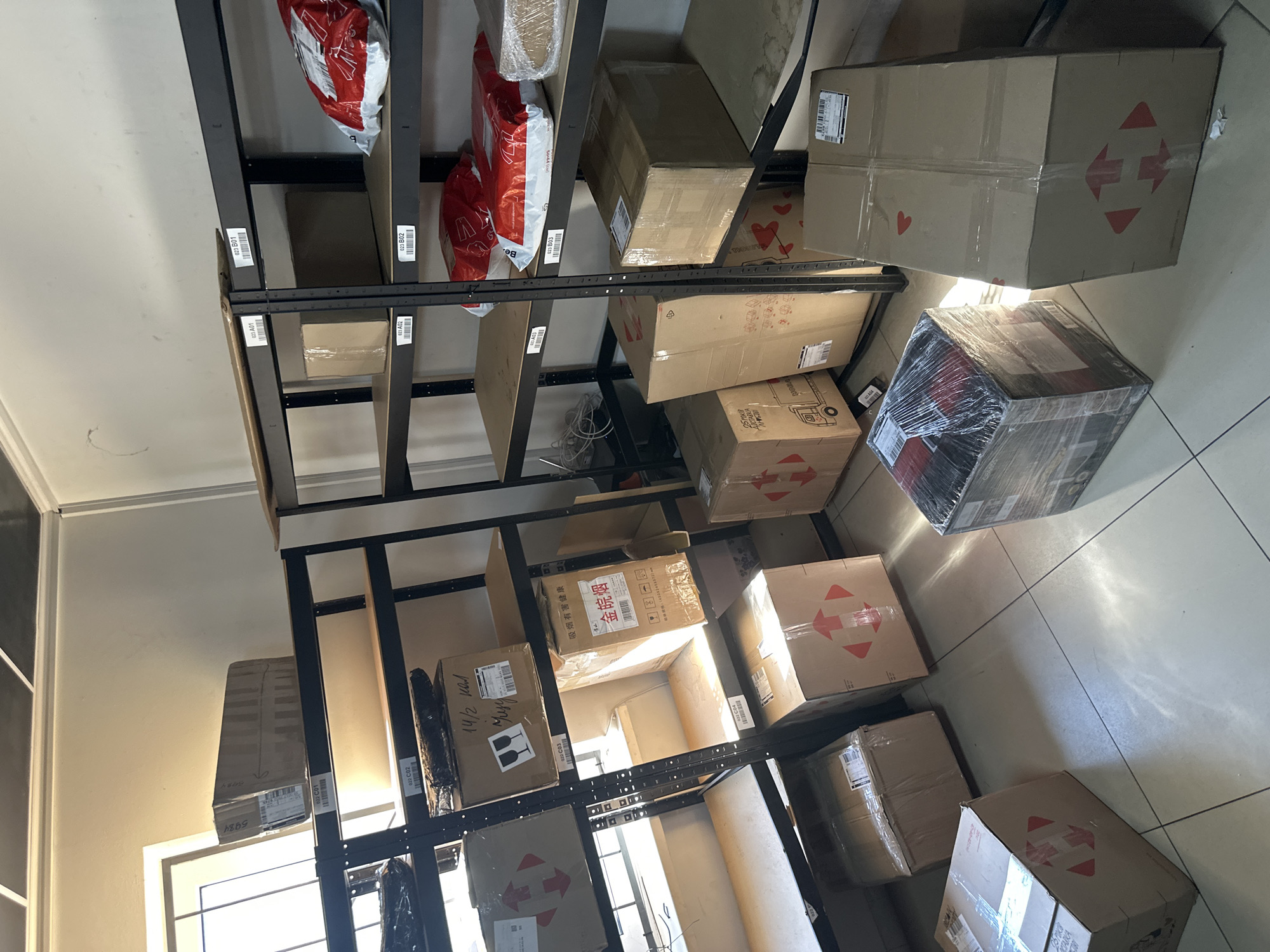 The image depicts the interior of a storage room, likely a fulfillment center or a retail backroom, in Khmilnyk, Ukraine. The room is organized with metal shelving units filled with various cardboard boxes of different sizes, some wrapped in plastic. On the shelves, there are also red and white packaged items, possibly mail or retail goods. The floor is tiled, and a window is visible on the left side of the image, letting in natural light. There are no people present in the scene. The overall impression is one of a functional, organized space for storing inventory.