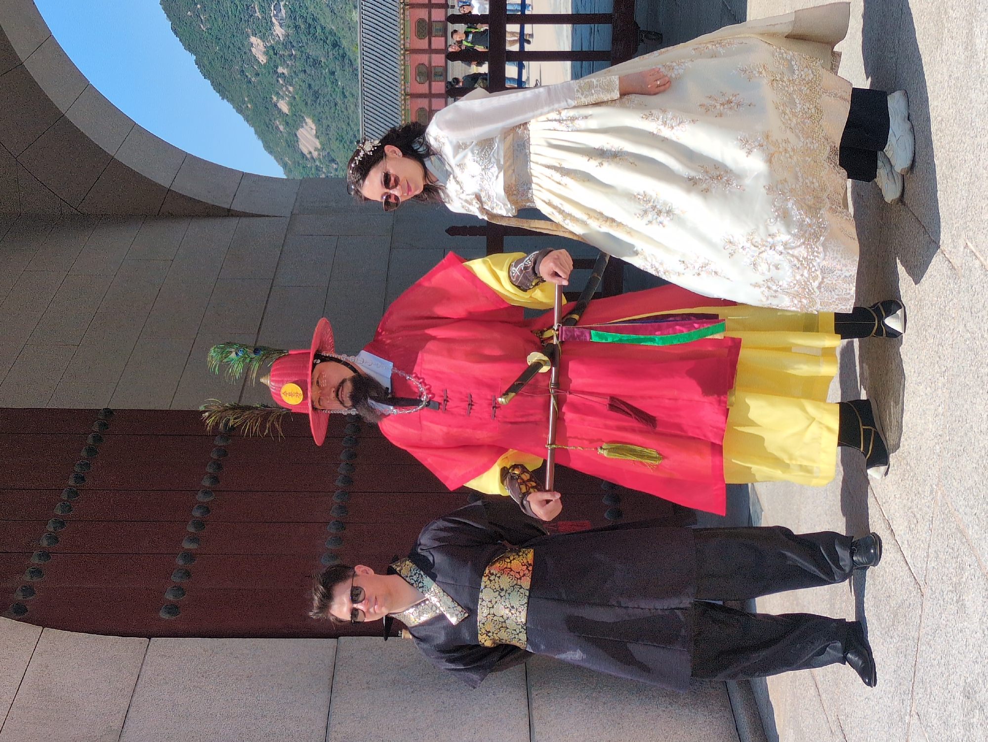 Three people are posing outdoors, near a building. The first person, a woman, is wearing a long white and gold Hanbok dress and sunglasses. The second person, a man with a beard, is wearing a red robe with a yellow underlayer, a red hat with a feather, and holding a sword. The third person, a man, is wearing a black robe with gold trim and sunglasses. The people are standing on a stone surface. The building has a wooden door with metal studs and a curved stone roof. In the background, there are trees, mountains, and other people. The location is likely in Seoul, South Korea, given the traditional Korean clothing.
