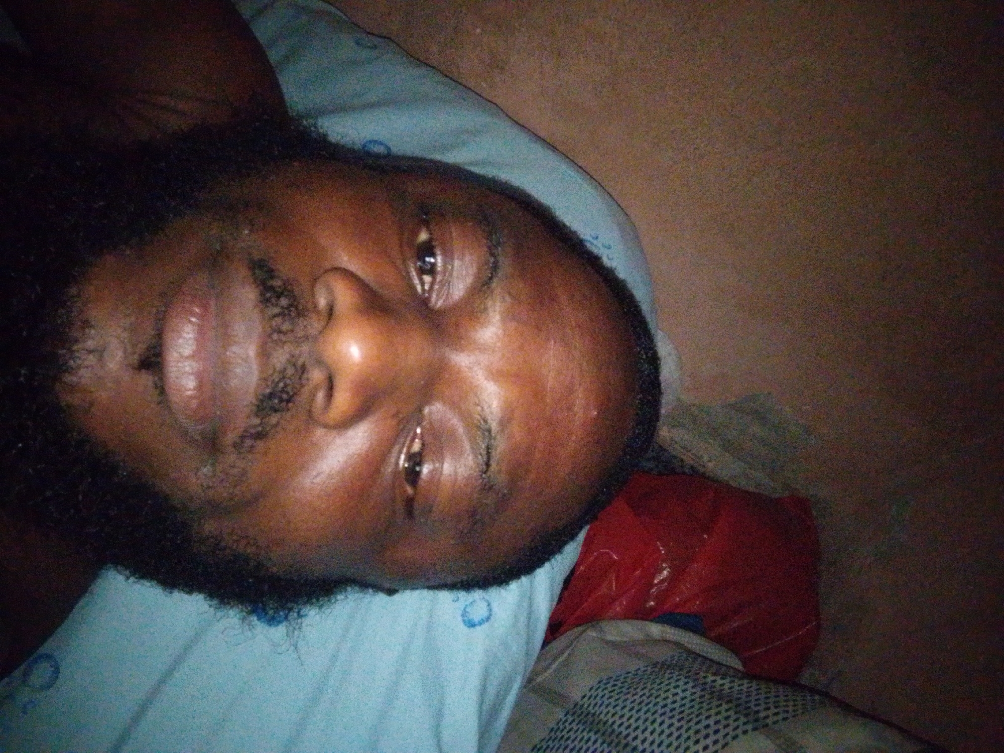 A close-up, low-angle shot of a man's face. He is lying down on a light blue pillow. The man has dark skin, a beard, and a mustache. His eyes are open, and he appears to be looking upwards. To the right of the pillow is a red plastic bag and a patterned sheet. The background is a plain, dark brown wall. The image appears to be taken indoors. There is no specific evidence to indicate the location as Ajaokuta, Nigeria.