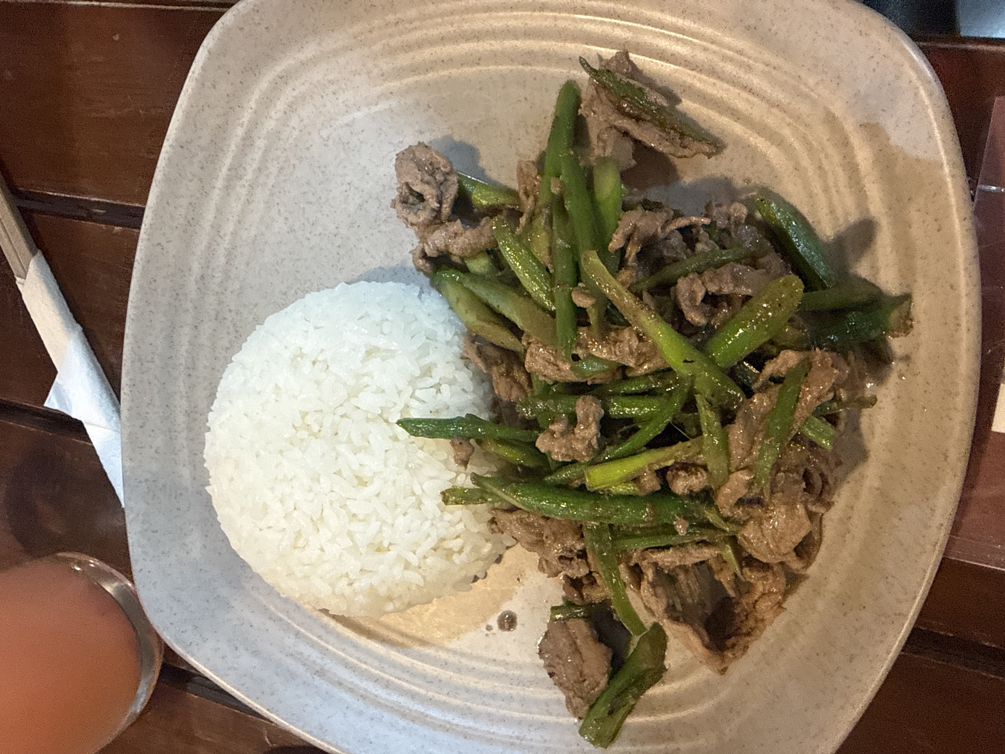The image shows a plate of food on a wooden table. The plate contains a mound of white rice and a portion of stir-fried beef with green beans. A glass of orange liquid is visible in the lower left corner of the frame. The location is identified as Mexico City, Mexico.