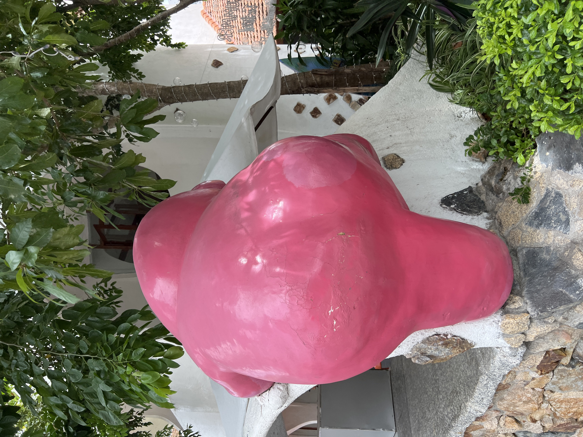 An outdoor scene in Nha Trang, Vietnam, features a large, bright pink, abstract sculpture positioned in the foreground. The sculpture has a bulbous, rounded shape, with a glossy, somewhat weathered surface. It is mounted on a base constructed from rough-hewn stones and concrete. Behind the sculpture, a white, multi-level building with arched openings is visible, partially obscured by lush green trees and foliage. A string of small, clear spherical decorations is wrapped around the trunk of one of the trees, and a sign with text is affixed to the building to the right. The environment appears to be a garden or courtyard area, with various plants and greenery surrounding the sculpture and building. The lighting suggests it is daytime.