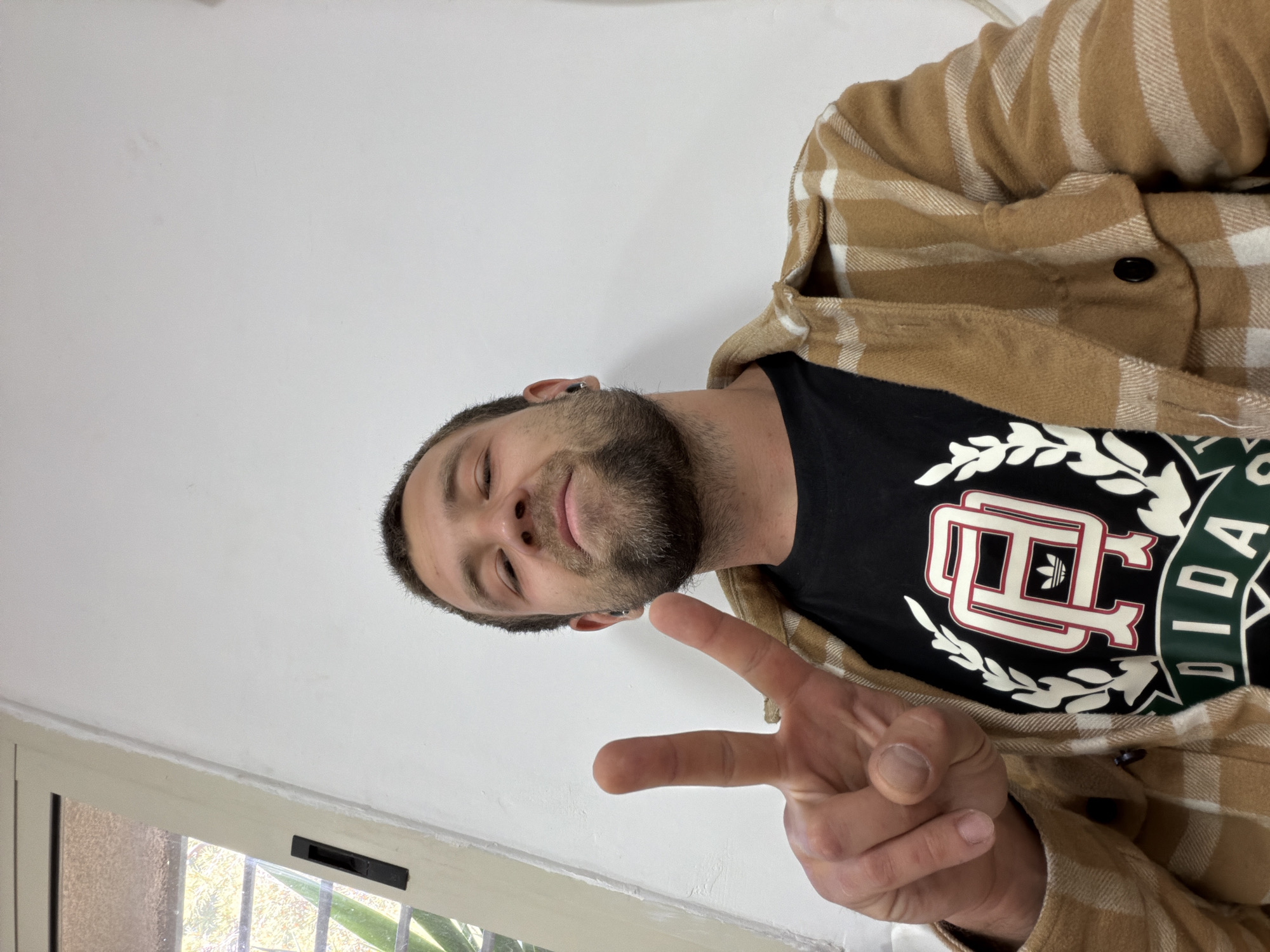 A man with short brown hair and a beard is indoors, facing downward towards the camera, giving a peace sign with his right hand. He wears a black t-shirt with a white, red, and green design, and a brown and white checkered shirt-jacket. The background is a plain white wall and a window with an obscured view to the outdoors. The location in the city is unknown.
