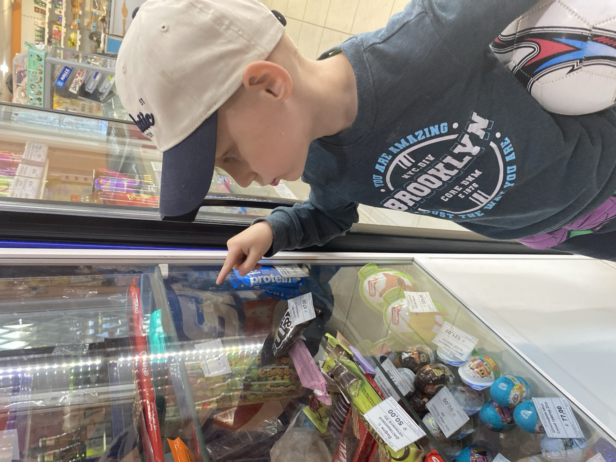 A young boy, wearing a white and navy baseball cap and a dark blue long-sleeved shirt, is seen leaning over a refrigerated display case in what appears to be a grocery or convenience store in Noviy, Russia. He is partially visible from the chest up, with a white and black soccer ball tucked under his left arm. The boy is looking intently into the case and pointing his right index finger at an item inside. His expression suggests curiosity or selection.
The display case contains various packaged snacks, chocolates, and candies. Visible items include a blue "protein" bar, several wrapped chocolate eggs, and other confectioneries. Price tags in Russian Rubles ("P") are attached to the products, showing amounts like "P 87,00," "P 50,00," "P 52,00," "P 77,00," and "P 121,00." Specific items mentioned on the labels are "Вафли с фисташковой начинкой 30г" (Wafers with pistachio filling 30g), "Шок. Яйцо Лошадки 20г" (Choc. Egg Horses 20g), "Шоколадное яйцо Синий трактор 20гр" (Chocolate egg Blue Tractor 20g), and "Десерт с подарком QUEST EGG 20" (Dessert with gift QUEST EGG 20). Other faint text includes "Calatali", "CHATOUN", and "TUKKO".
In the background, shelves are visible with products, including small boxes labeled "HALLS," "birol," and "Orbit." The boy's shirt features a circular graphic with text that reads: "YOU ARE AMAZING BROOKLYN NYC. DIV CORE DNM E 1975." The scene captures a moment of a child choosing a treat in a retail setting.