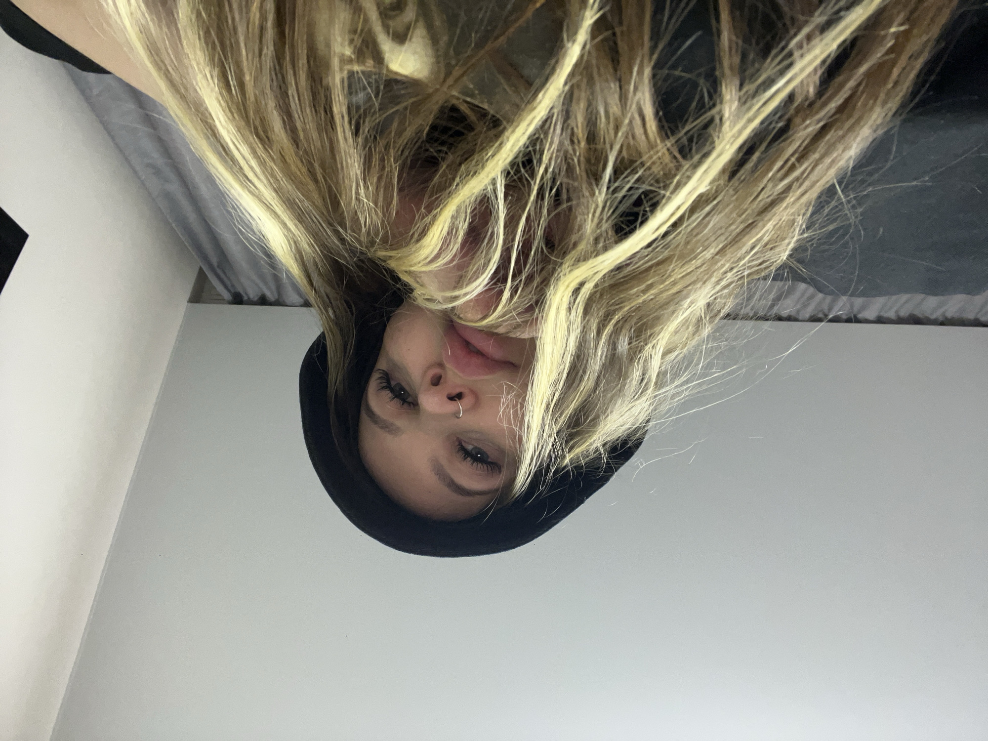 The image presents a close-up, upside-down view of a young woman, likely taken from above her while she is in a reclined position. Her face is visible in the upper-middle section of the frame, framed by her long, blonde hair that appears to be flowing downwards (towards the top of the image in this orientation). She is wearing a dark, possibly black, hooded sweatshirt, with the hood framing her face.
Her features include well-defined eyebrows and noticeable eyelashes, suggesting makeup. She has a small, silver-colored nose ring in her right nostril. Her lips are slightly parted, and her eyes are open, looking directly towards the camera. Her hair is a mix of blonde and lighter brown tones, appearing somewhat tousled and natural.
The background behind her head is dark and indistinct, possibly a bed or pillow. The majority of the lower half of the image (below her face in this orientation) consists of a plain, light-colored surface, which could be a wall or ceiling.
There is no explicit activity beyond posing for the camera. The image conveys a casual and relaxed mood. No specific time of day or weather conditions can be inferred. No text is visible in the image. While the provided context indicates Krasnodar, Russia, there are no visual cues within the image to confirm this location.