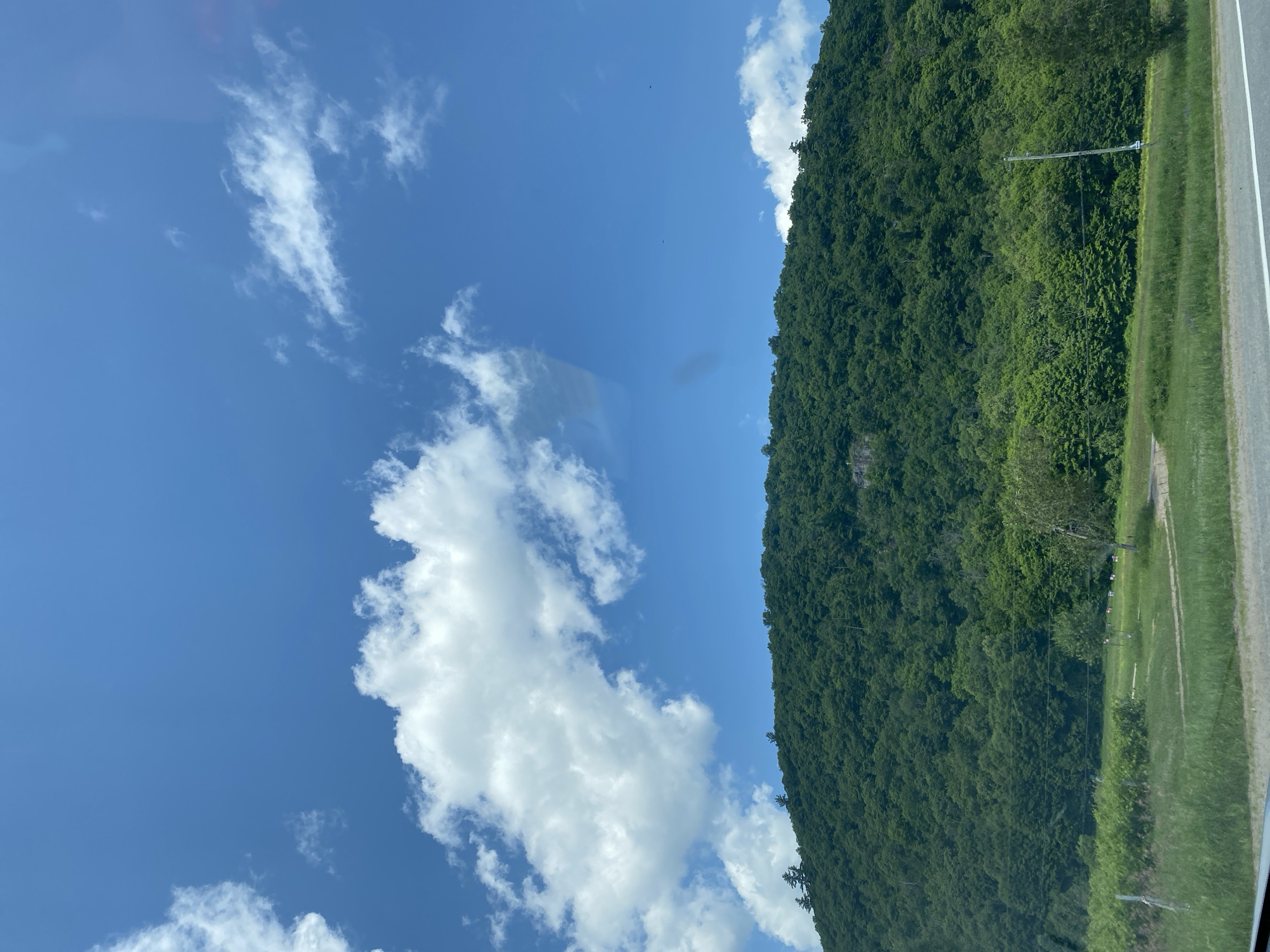 The image depicts a vast, green, forested hillside under a bright blue sky dotted with fluffy white clouds. The hillside occupies the majority of the frame, with its dense foliage creating a lush, textured surface. At the bottom of the image, a grassy field stretches towards the hillside, with a paved road visible in the foreground. The scene appears to be a natural landscape, possibly a park or a rural area. The presence of a road suggests it may be a scenic route or a view accessible from a roadway. There are no visible people or structures on the hillside. The overall impression is one of natural beauty and tranquility. The location of Ripon, Canada, is not directly evident from the visual elements of the image itself; this information would need to be provided contextually.