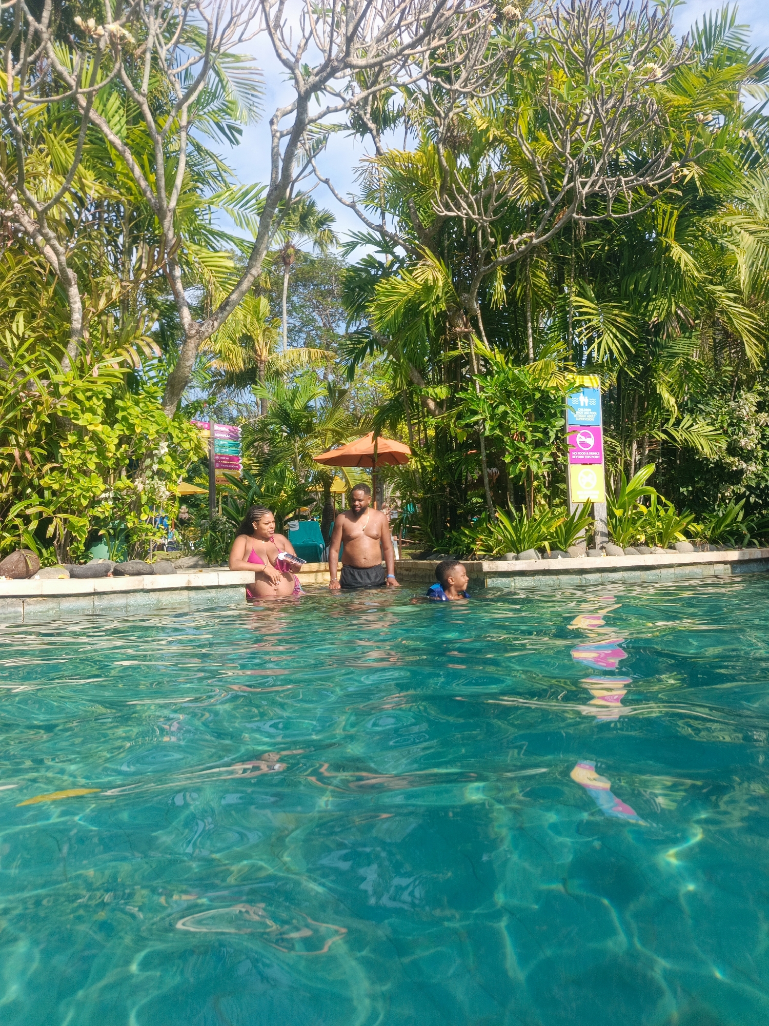 In a sun-drenched outdoor swimming pool, three individuals are present. A woman, appearing to be pregnant, stands near the pool's edge wearing a pink bikini. Next to her, a shirtless man in black swim trunks also stands in the water. To the left, a young boy wearing a life vest is swimming in the pool. The pool water is a vibrant turquoise color, with sunlight creating rippling patterns on the surface. Behind the pool, lush green tropical vegetation, including palm trees and other foliage, forms a dense backdrop. An orange umbrella is visible in the background, as are several informational signs. The overall setting suggests a resort or a leisure facility in a tropical location, likely Kuta, Indonesia, known for its beaches and resorts.