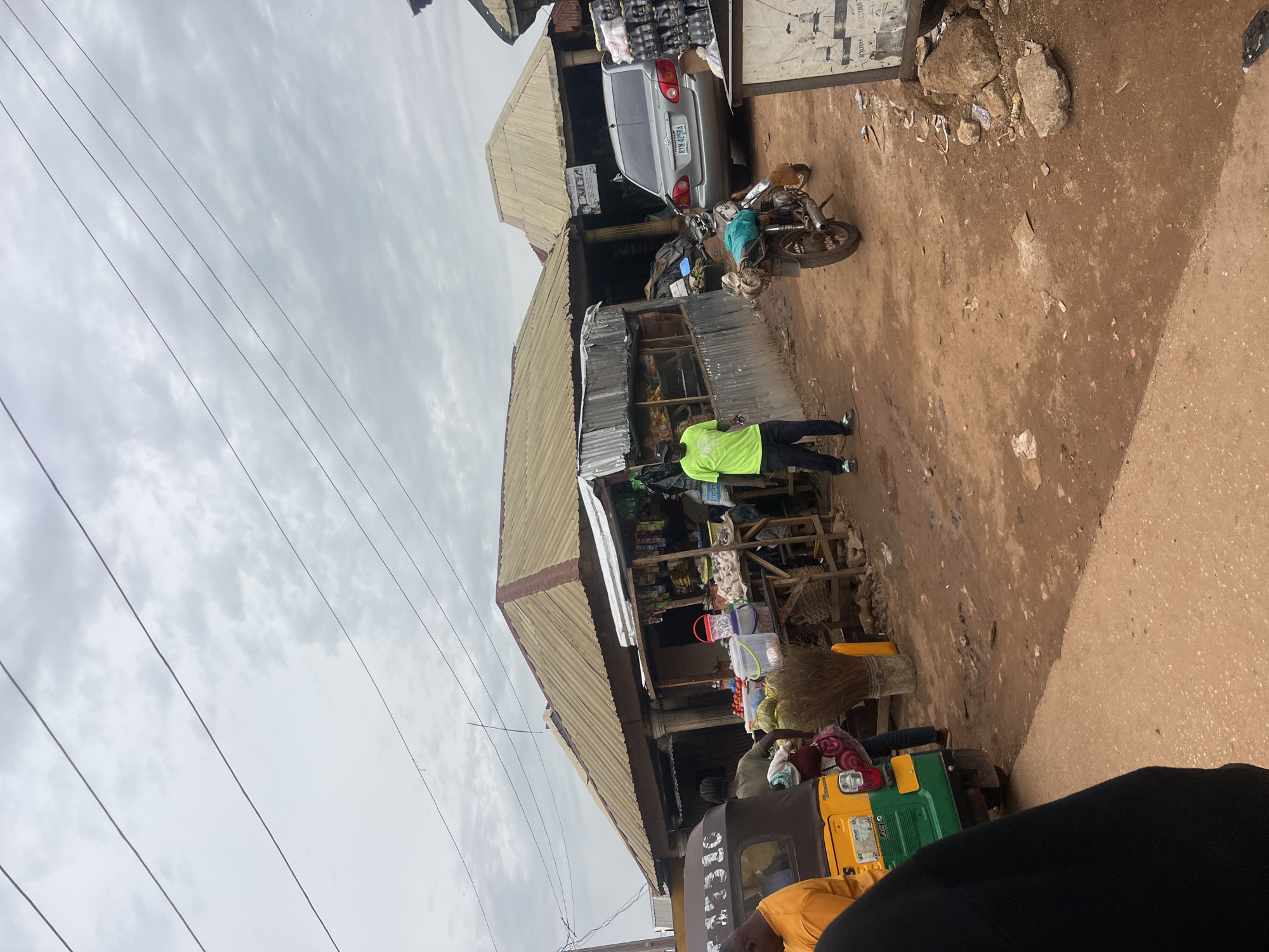 This image depicts a street scene in Bokkos, Nigeria, likely in a market area. The foreground shows a portion of a yellow and green auto-rickshaw. Behind it, several market stalls are visible, constructed with corrugated metal and wood, topped with weathered, tan-colored roofs. One stall displays fruits like tomatoes and gourds, along with buckets and a large broom.
A man wearing a bright green t-shirt and black pants stands near a stall, looking towards the viewer. Another individual, partially obscured, is visible near the auto-rickshaw. In the background, a silver sedan is parked, and a motorcycle with bags strapped to its rear is also present. The ground is unpaved, appearing to be dirt and mud. Power lines crisscross the overcast sky, which is filled with gray clouds. The overall impression is one of a bustling, everyday commercial environment in an African town.