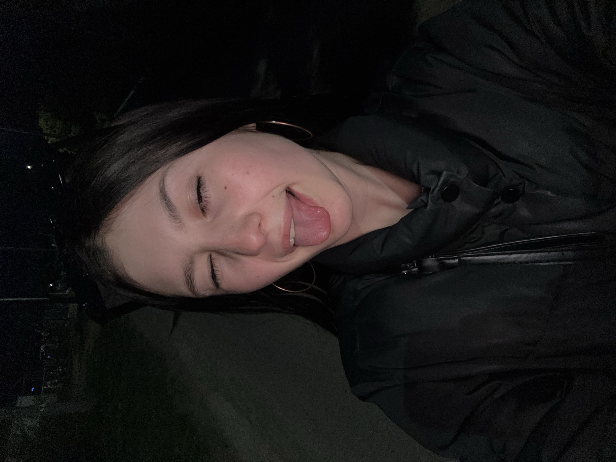 A woman is outdoors at night. She has her eyes closed, tongue out, and is wearing a black puffer jacket and large hoop earrings. The location appears to be Фоки, Russia.