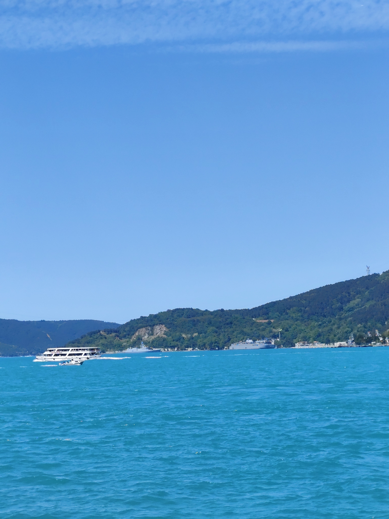 The image shows a daytime view of the Bosphorus in Sarıyer, Turkey. The scene includes turquoise water with gentle waves. A white passenger ferry is moving across the water from left to right, creating a white wake. In the background, green hills covered in trees are visible, partially obscured by the distance haze. A large gray warship is docked along the coastline in the distance, near small structures that are also likely houses. The clear blue sky has a few wispy clouds.
