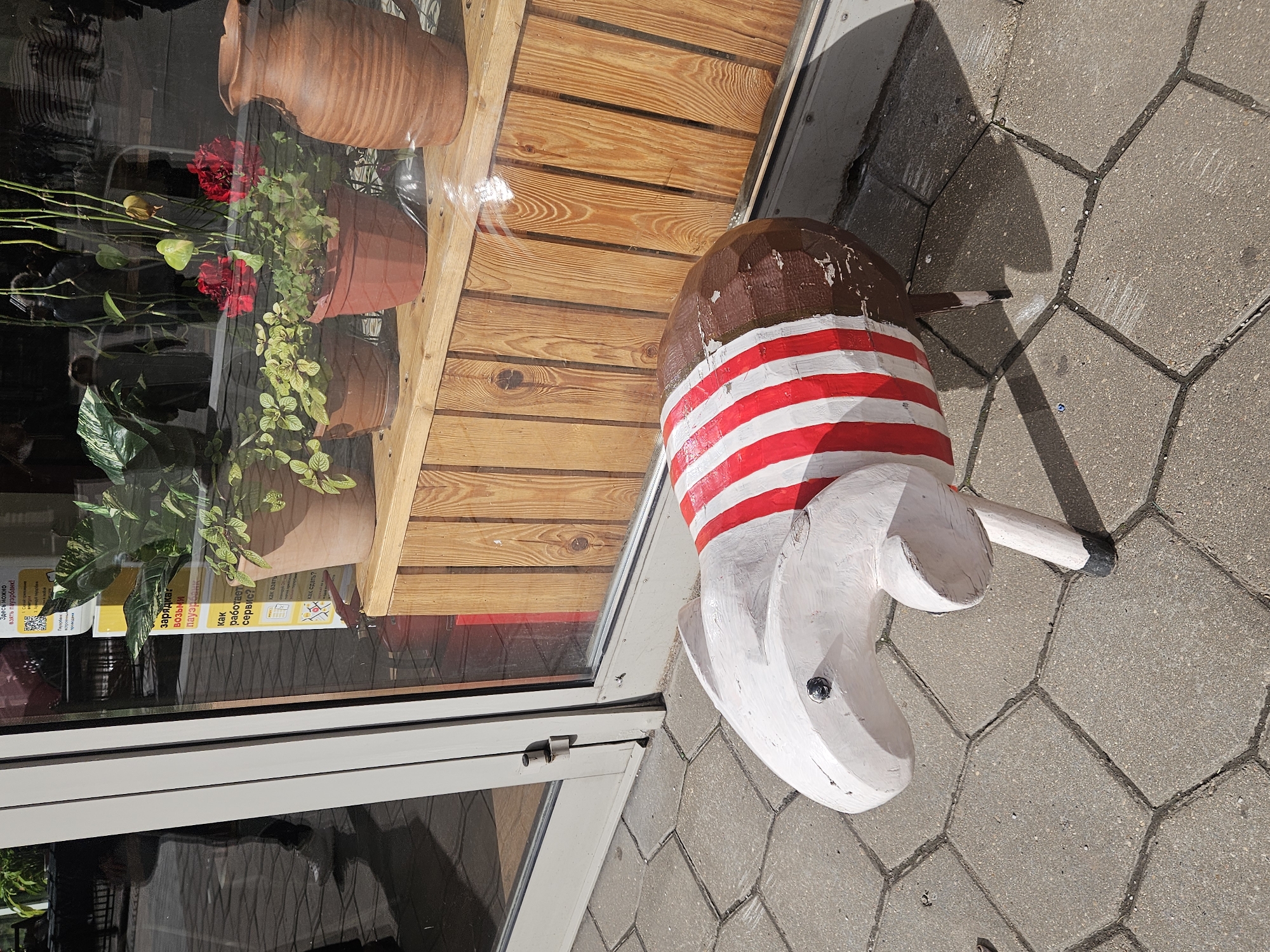 Here is a matter-of-fact description of the image:
A decorative wooden statue of an animal resembling a pig or anteater stands on a grey bricked surface, directly adjacent to a storefront window. The statue is painted in a rustic style. The head is white with a black dot for an eye, the body has a brown upper section and the midsection has a pattern of alternating red and white stripes.
The storefront window features a display of plants in terracotta pots placed on wooden shelves. The interior of the store is reflected in the window, creating a blurred effect. The window frame is light grey.
The image appears to be taken outdoors in daylight in the city of Unknown, Unknown.