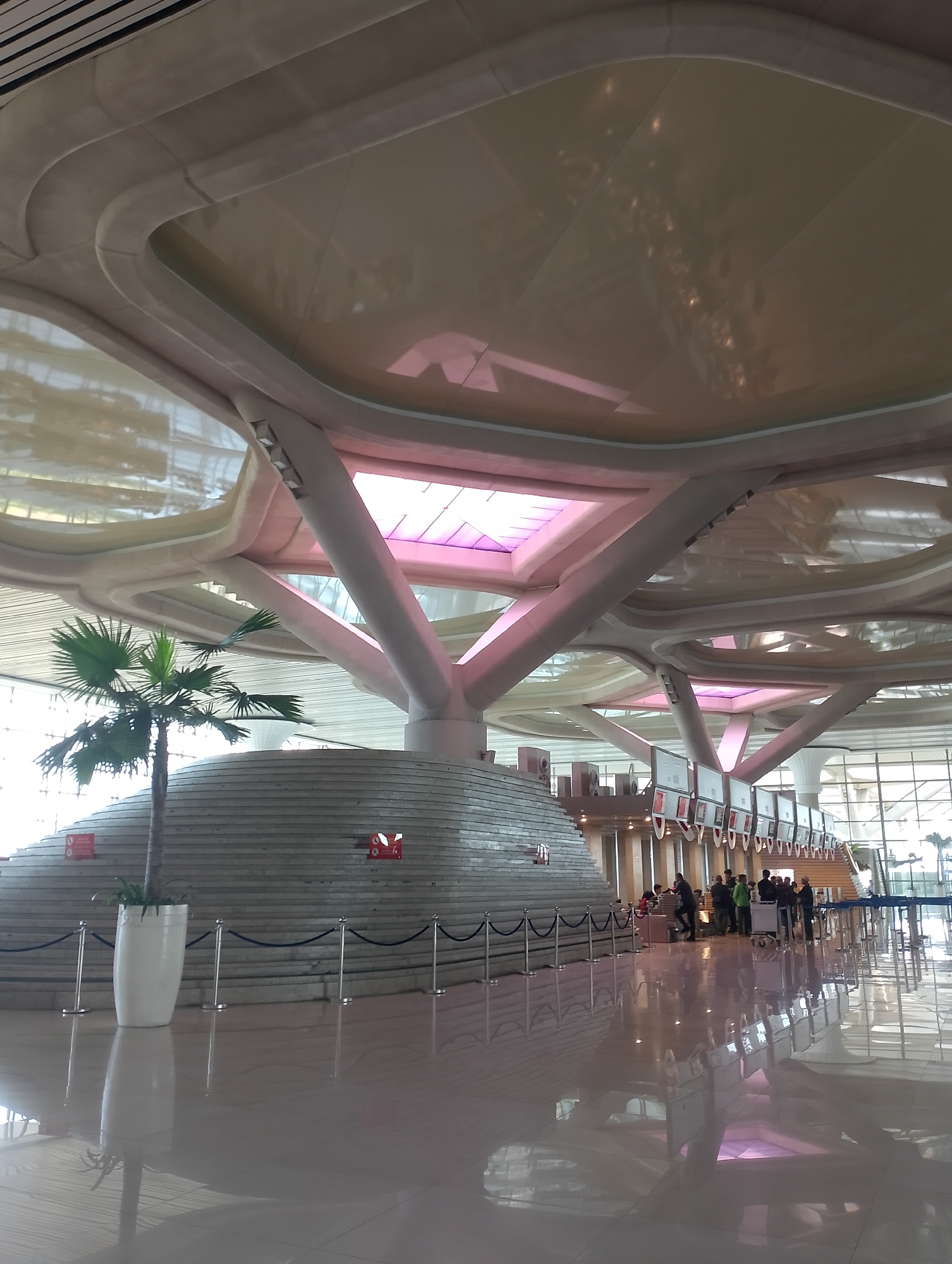 The image shows the interior of a modern airport terminal in Palihan, Indonesia. The ceiling is a complex structure of white, curved beams that support translucent panels, some of which emit a pinkish light. In the foreground, a polished, reflective floor shows the distorted reflections of the ceiling and structural elements. To the left, a large white planter contains a small palm tree. Behind the planter, a tiered, stepped structure made of gray stone is visible, with red warning signs affixed to its side. A metal stanchion and rope barrier runs across the lower part of the image. Further into the terminal, a group of people are gathered around check-in counters, with some queuing and others interacting with staff. Luggage carts are also present. The overall environment appears bright and spacious, with large windows in the background indicating a connection to the outdoors.