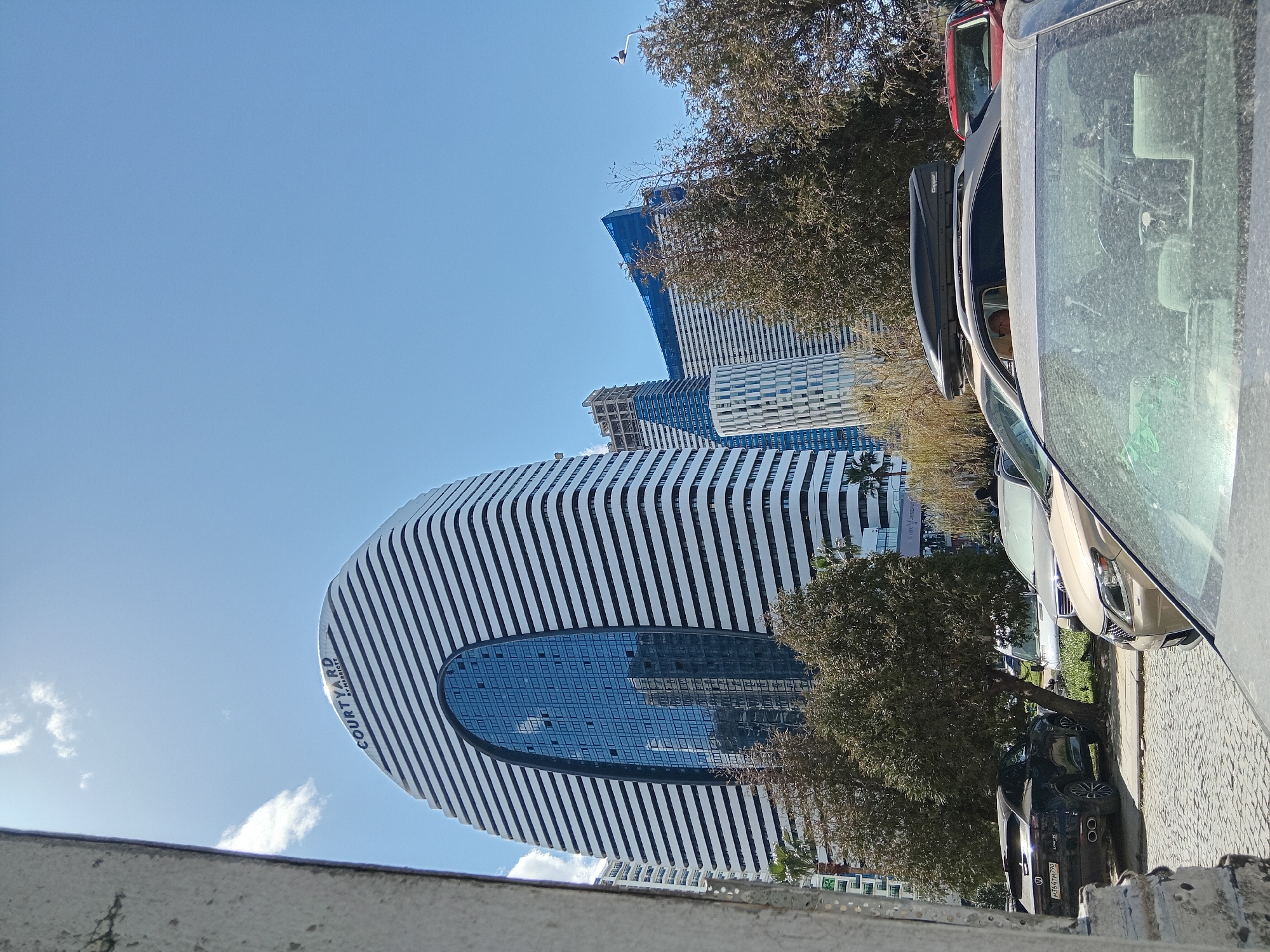 The image shows a view of the Courtyard by Marriott hotel in Batumi, Georgia, from a street-level perspective. The hotel has a distinctive oval shape with horizontal white stripes and a large elliptical window facade. Adjacent to the hotel are other high-rise buildings, some of which appear to be under construction, covered in blue protective material.
Parked cars line the right side of the frame, including a dark-colored SUV and a silver car. In the lower left corner is a view of a low concrete wall. Trees partially obscure the view of the hotel and surrounding structures. The sky is clear and blue with some clouds.