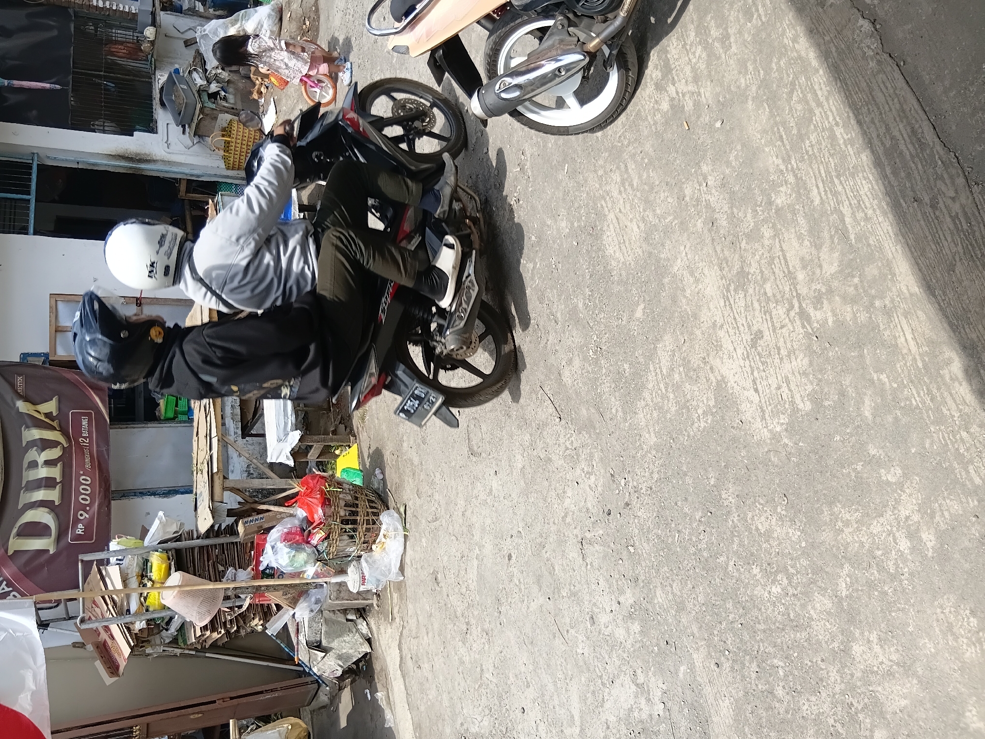 The image depicts a street scene in Tanggul Wetan, Indonesia, likely on a sunny day. In the foreground, a dark-colored motorcycle is partially visible on the right side of the frame, with its exhaust pipe and wheel in clear view. The motorcycle is parked on a concrete surface that appears worn and textured, with visible cracks and debris scattered across it.
The main focus of the image is a two-person motorcycle traveling away from the viewer. The rider and passenger are both wearing helmets, the rider in black and the passenger in white. They are on a black and red motorcycle. The passenger is wearing a gray jacket and dark pants, while the rider's attire is mostly obscured. Their legs are visible, with the passenger wearing white sandals over dark socks.
Behind the moving motorcycle, a young girl in a light-colored dress and pink shorts is visible on the right, facing away from the camera. She appears to be standing next to a small pink bicycle.
Further in the background, a building with a light-colored facade is present. There is a sign above an entrance that reads "DIRIA" in large, brown letters, with smaller text below indicating "RP 9.000 /BUNGKUS(12 BATANG)". To the left of the building, a pile of cardboard boxes and other items are stacked against a metal frame, some of which are contained in plastic bags. A woven basket filled with items, including red plastic bags, is also visible. The overall environment suggests a busy, possibly commercial area with elements of both everyday life and a slightly unkempt appearance.