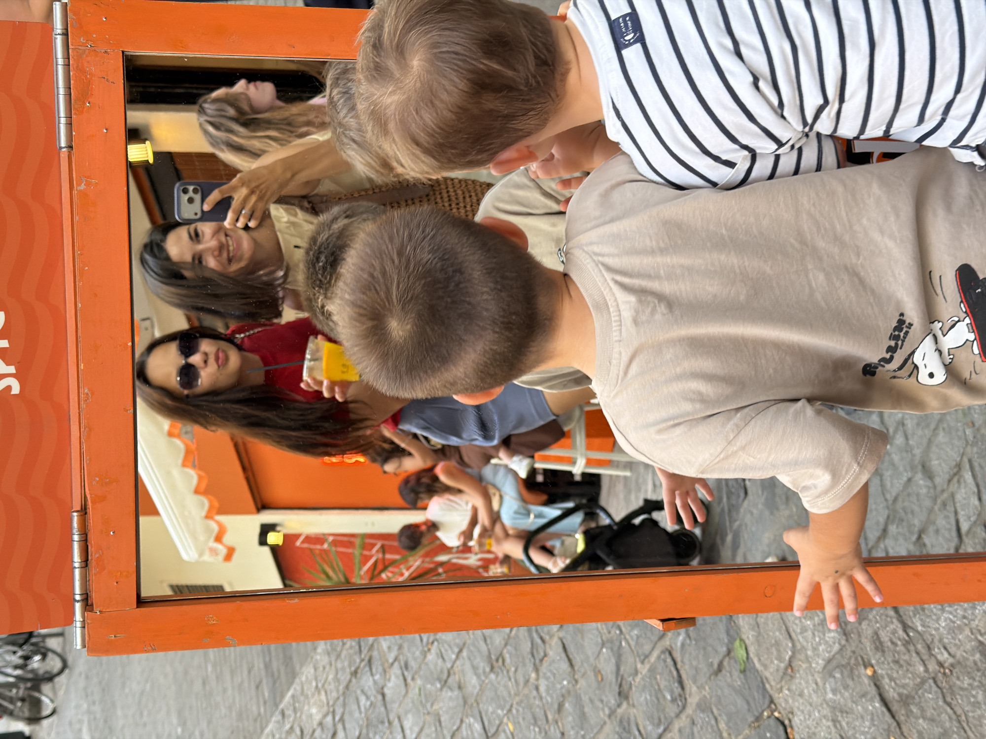 In an outdoor urban setting, two young boys are looking into a large, orange-framed mirror. The boy on the left wears a light brown t-shirt with a Snoopy graphic. The boy on the right wears a black and white striped t-shirt. Their backs are to the camera as they face the mirror, which reflects a street scene. In the reflection, three women are visible. One woman in the foreground of the reflection is wearing sunglasses and a red shirt, holding a yellow cup. Another woman next to her is smiling and taking a selfie with a phone. A third woman with blonde hair is partially visible to the right. Behind them, in the reflection, are other people seated at tables, an orange awning, and a brick-paved ground. A black stroller is also visible in the reflection, near the base of the mirror.