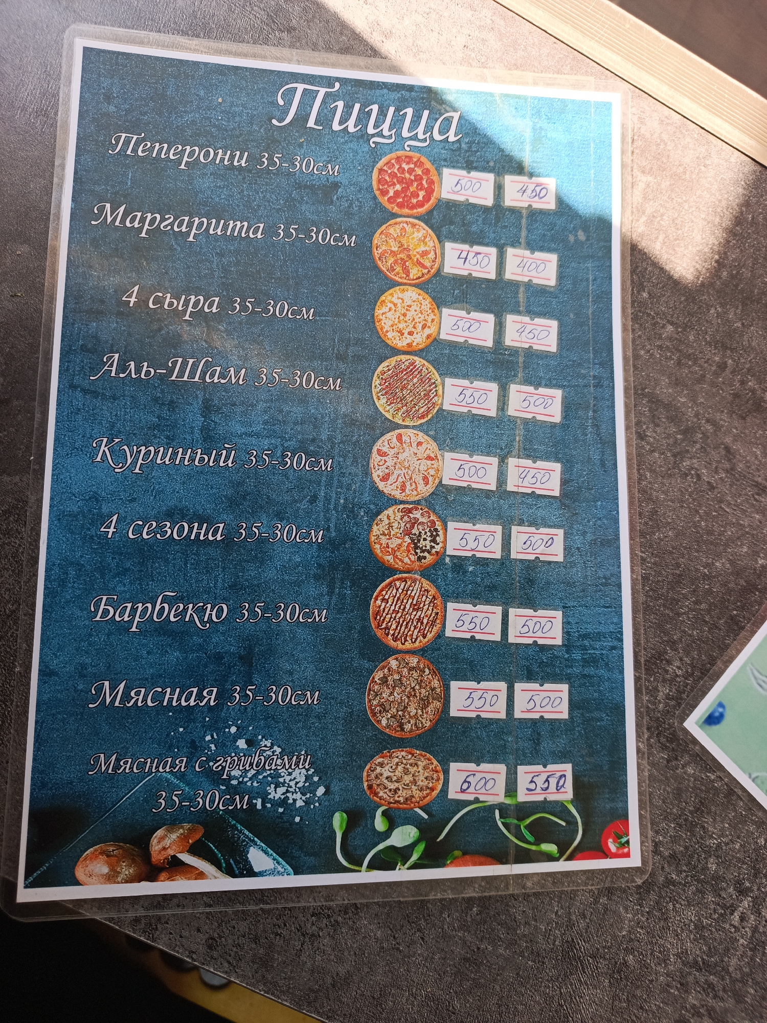 The image shows a menu for pizza displayed on a textured blue background, likely in a restaurant or food establishment. The menu lists various pizzas with their names and sizes in centimeters, along with prices. Some of the pizzas listed are "Пеперони" (Pepperoni), "Маргарита" (Margherita), "4 сыра" (4 Cheeses), "Аль-Шам" (Al-Sham), "Куриный" (Chicken), "4 сезона" (4 Seasons), "Барбекю" (Barbecue), "Мясная" (Meat), and "Мясная с грибами" (Meat with Mushrooms). Each pizza is accompanied by an image and two price tags. The prices are handwritten on small white labels. The bottom of the menu features illustrations of mushrooms, sprouts, and tomatoes. The menu is encased in clear plastic, suggesting it is a physical menu designed to be durable. The overall presentation indicates a casual dining setting. There is no visible indication or information related to the city of Üç-Korgon, Kyrgyzstan, in the image.