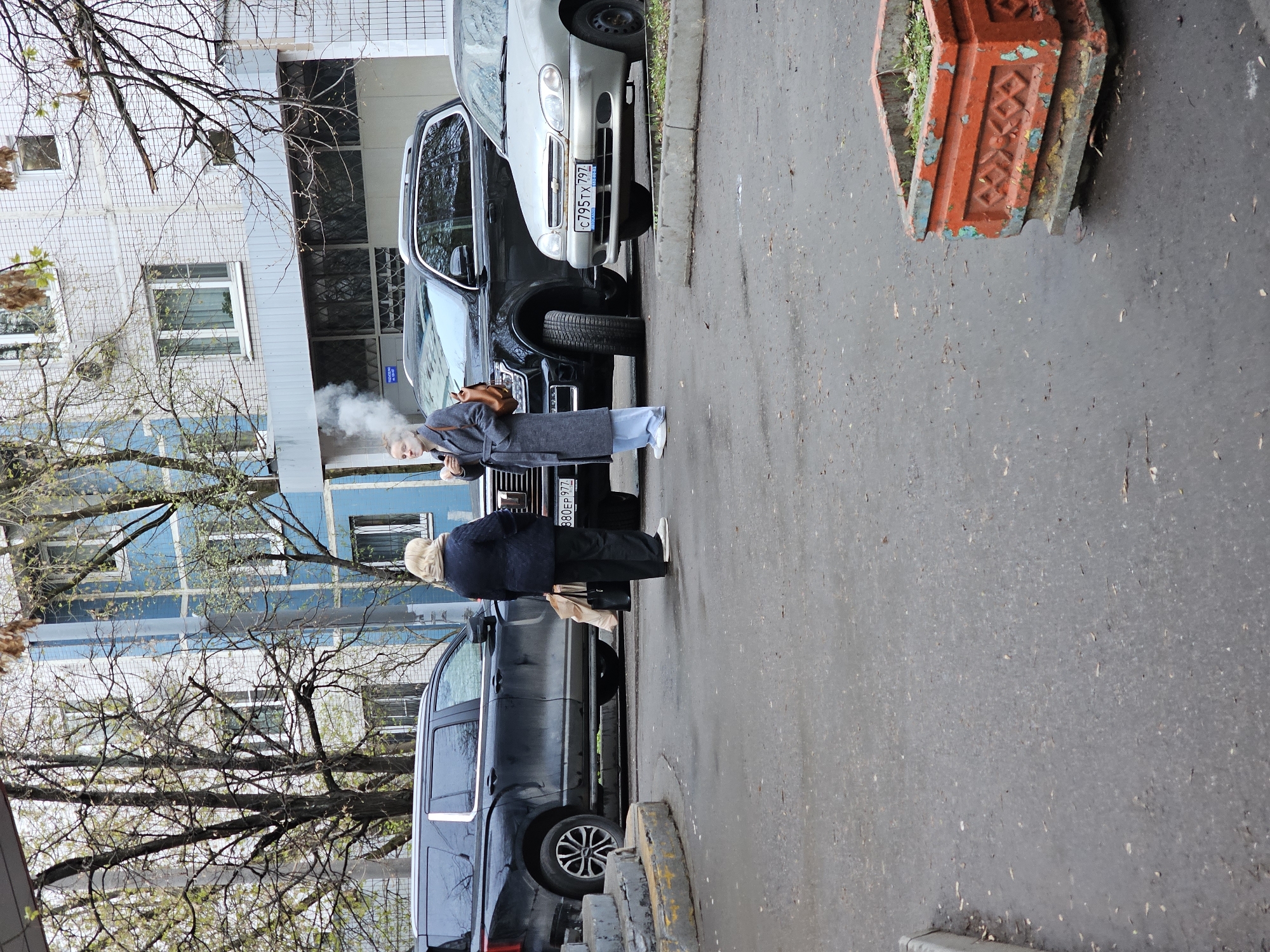The image depicts an outdoor urban or suburban scene in Khimki, Russia, likely during late winter or early spring due to the bare trees and muted lighting. Two individuals are featured near parked cars on an asphalt surface, with a residential building in the background.
A younger woman, dressed in a grey long coat, light blue jeans, and white sneakers, is visible from the side. She is actively vaping, with a distinct cloud of vapor emanating from her mouth. She carries a brown leather satchel over her shoulder. Adjacent to her, with their back to the camera, stands an older person wearing a dark coat and trousers, appearing to be engaged in conversation or interaction with the younger woman.
Several cars are parked along the side of the street. A silver car (possibly a Chevrolet Lanos) is prominently displayed with the Russian license plate C795TX 797, indicating the Moscow region. Further down, a dark SUV or minivan is visible, bearing the license plate 380EP 977, also from the Moscow region.
The background features a multi-story building with a light-colored, possibly tiled, facade and multiple windows. The building's architectural style appears typical of post-Soviet era residential structures. Bare tree branches, characteristic of the season, are spread across the upper part of the frame, partially obscuring the building. An old, weathered red concrete planter with a geometric pattern is situated on the right side of the asphalt. The overall scene captures an ordinary street moment.