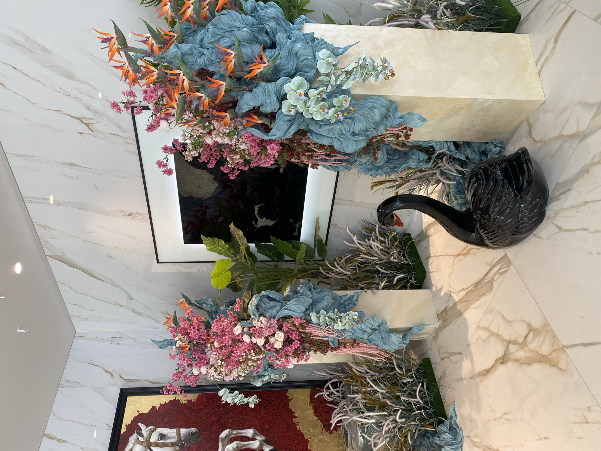 This indoor scene shows a decorated area with two white, rectangular pedestals. Each pedestal is adorned with arrangements featuring orange bird-of-paradise flowers, pink and white blossoms, blue fabric resembling waves, and light green orchids. One pedestal is placed beside a framed painting with a red and gold background depicting white horses, while the other is beside a framed abstract black and white image. A black swan statue is positioned near the upper pedestal. The background consists of marble-like tiled walls and floors. The location appears to be indoors, but there is no information to confirm that it is indeed in Sochi, Russia.