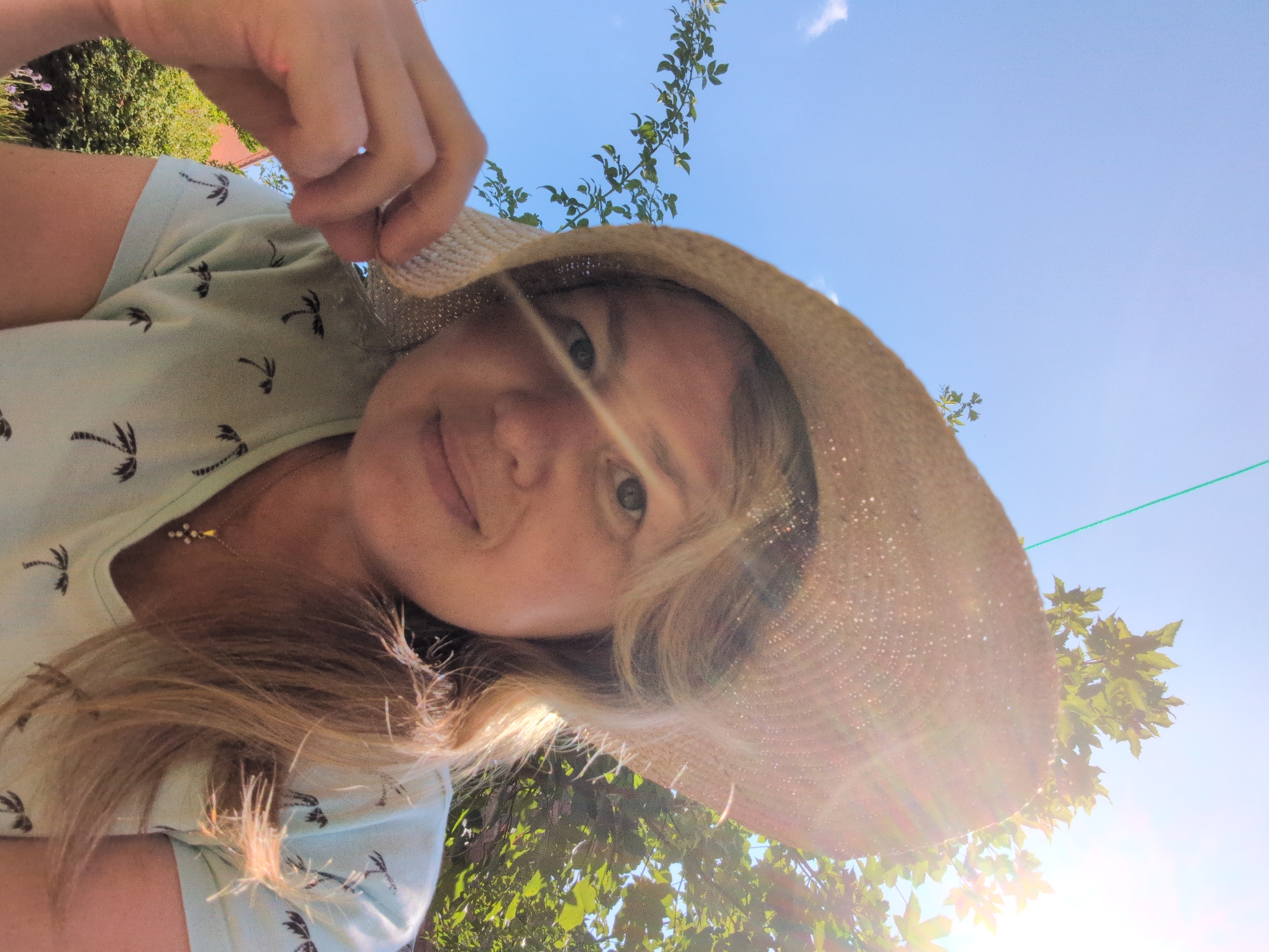 A woman wearing a straw hat and a light blue t-shirt with a palm tree pattern is shown from a low angle, looking up at the camera. She is outdoors, with a bright blue sky and green foliage in the background. The sun is shining brightly, creating a lens flare effect. Her left hand is touching the brim of her hat. She is wearing a small gold necklace with a cross pendant. The image appears to be a selfie. The location is not specified in the image or its context.