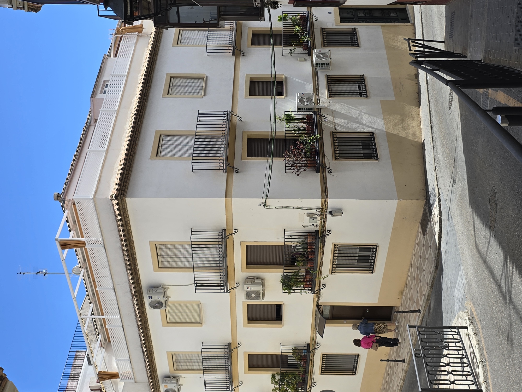 The image shows a corner view of a multi-story building with a white facade and a yellowish-ochre base on the lower levels. The building has several balconies with black metal railings, some adorned with potted plants. Multiple windows are visible, some with blinds and others with security bars. Air conditioning units are mounted on the exterior walls. On the rooftop, there is a white structure with a lattice railing and tan fabric awnings.
Two women are standing on the sidewalk in front of the building, near a doorway. One woman, wearing a pink jacket and dark pants, carries a beige handbag. The other woman wears a denim jacket and a patterned skirt. They appear to be interacting near the entrance of the building.
The building is situated on a street with a paved surface and a sidewalk made of patterned tiles. Black metal railings line the edge of the sidewalk in the foreground. The sky is clear and blue. The scene suggests a sunny day in an urban environment. Based on the architectural style, which is typical of Mediterranean regions, and the presence of the olive tree motif on the balconies and rooftop railings, this location is likely in the city of Jaén, Spain.