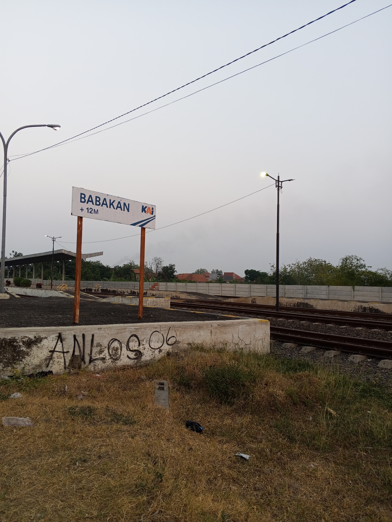 The image depicts a railway station signage at Babakan, +12M, identified by the KAI logo, indicating it is likely a station under the Indonesian railway operator, PT Kereta Api Indonesia. The sign is mounted on two wooden poles in front of a platform with dark ballast. To the left, a street lamp is visible, and further back, a covered platform structure with pillars is present. The environment consists of dry grass, overgrown vegetation, and concrete barriers along the railway tracks. Graffiti, including "ANLOS 06," is scrawled on a concrete wall in the foreground. The sky is overcast, suggesting a potentially early morning or late afternoon setting. The general scene is one of a functional but somewhat neglected railway infrastructure.
