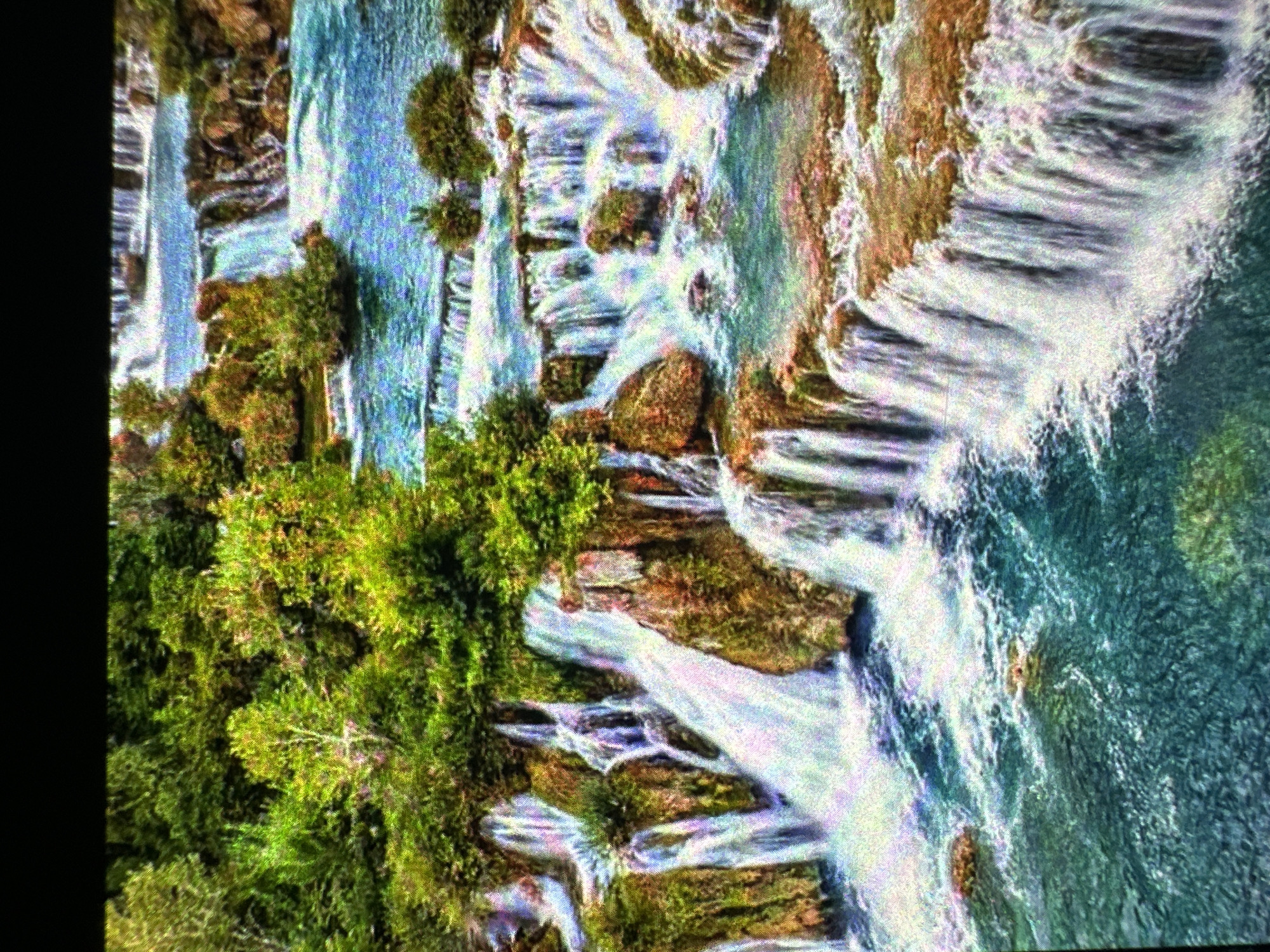A cascading waterfall system is depicted, with lush green trees and vegetation growing from rocky outcrops surrounding the various water levels. The water flows over tiered rock formations, creating a dynamic and natural landscape. The color palette is dominated by vibrant greens of the foliage and the striking blues and whites of the water and foam, interspersed with the earthy tones of the rocks. The scene suggests a pristine, natural environment, possibly a national park or a wilderness area. The image is captured from a slightly elevated perspective, offering a panoramic view of the falls and their surroundings. The lighting appears to be natural daylight, highlighting the textures and details of both the water and the plant life.